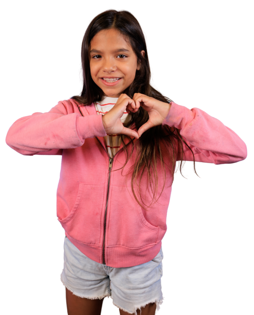 A young girl with long dark hair smiling and making a heart shape with her hands, wearing a pink hoodie and gray shorts.