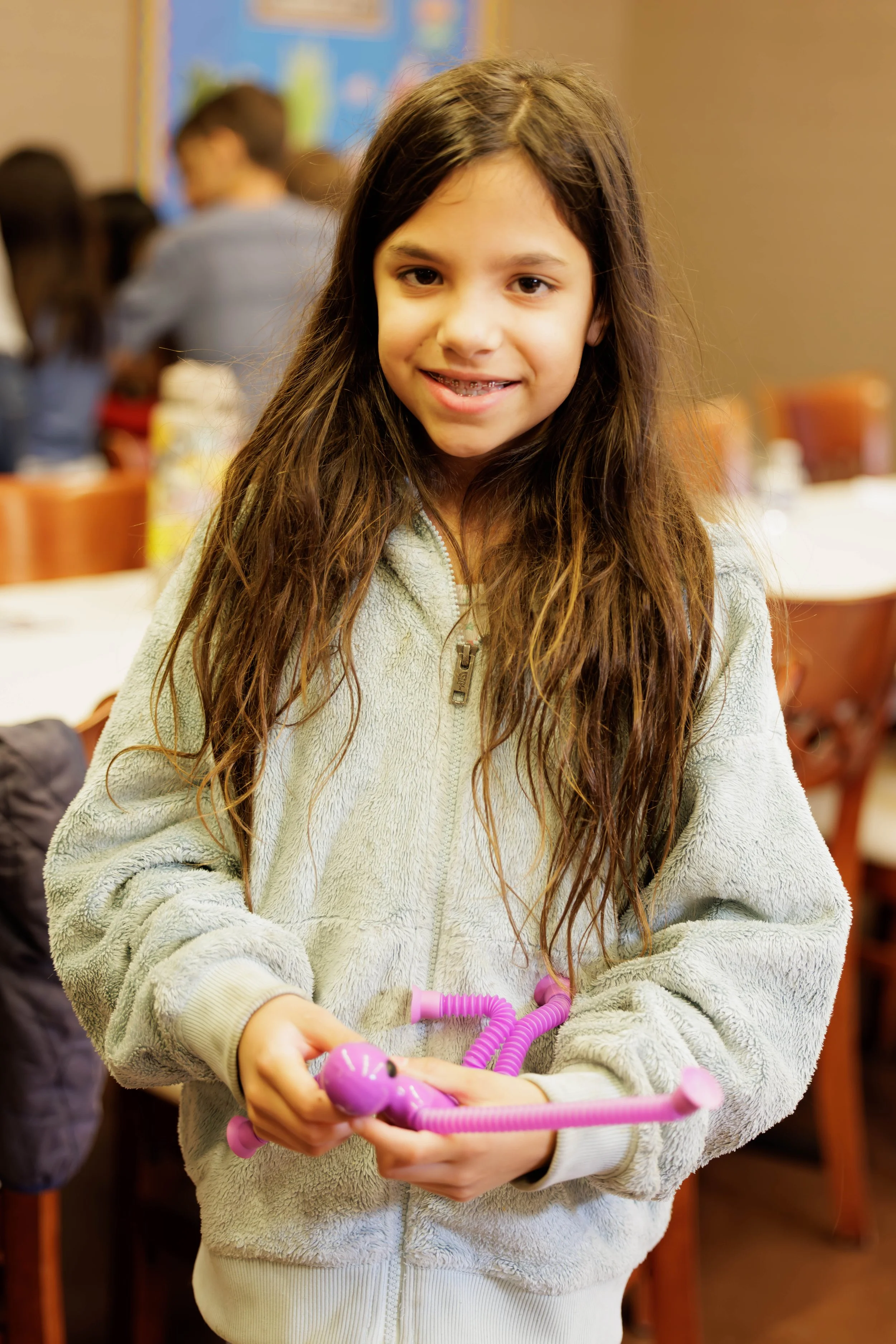 A young girl with long brown hair and a gap-tooth smile, wearing a light gray fleece jacket, holding a pink plastic toy in her hands, standing in a room with other children and tables in the background.
