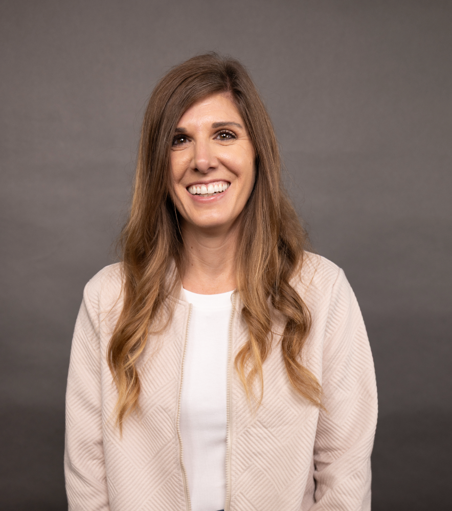 A woman smiling with long, wavy brown hair, wearing a light-colored jacket and white top, standing against a plain gray background.