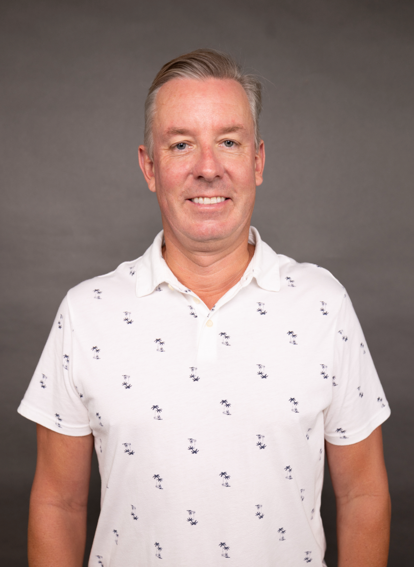 A man with blonde hair and a light complexion smiling, wearing a white polo shirt with small palm tree patterns, against a gray background.