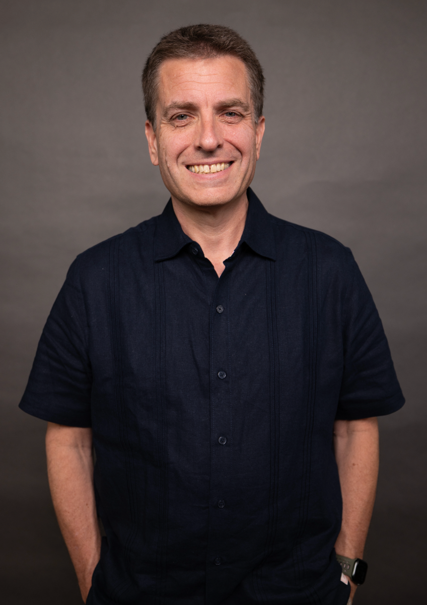 A middle-aged man with short brown hair, smiling with a closed mouth, dressed in a black collared shirt, standing against a neutral gray background.