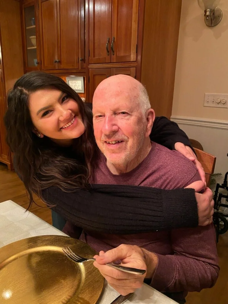 A young woman hugging an older man, both smiling, at a dinner table in a home kitchen with wooden cabinets in the background.
