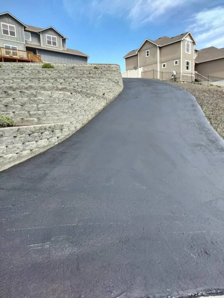A freshly paved asphalt driveway on a hill leads up to suburban houses under a blue sky.