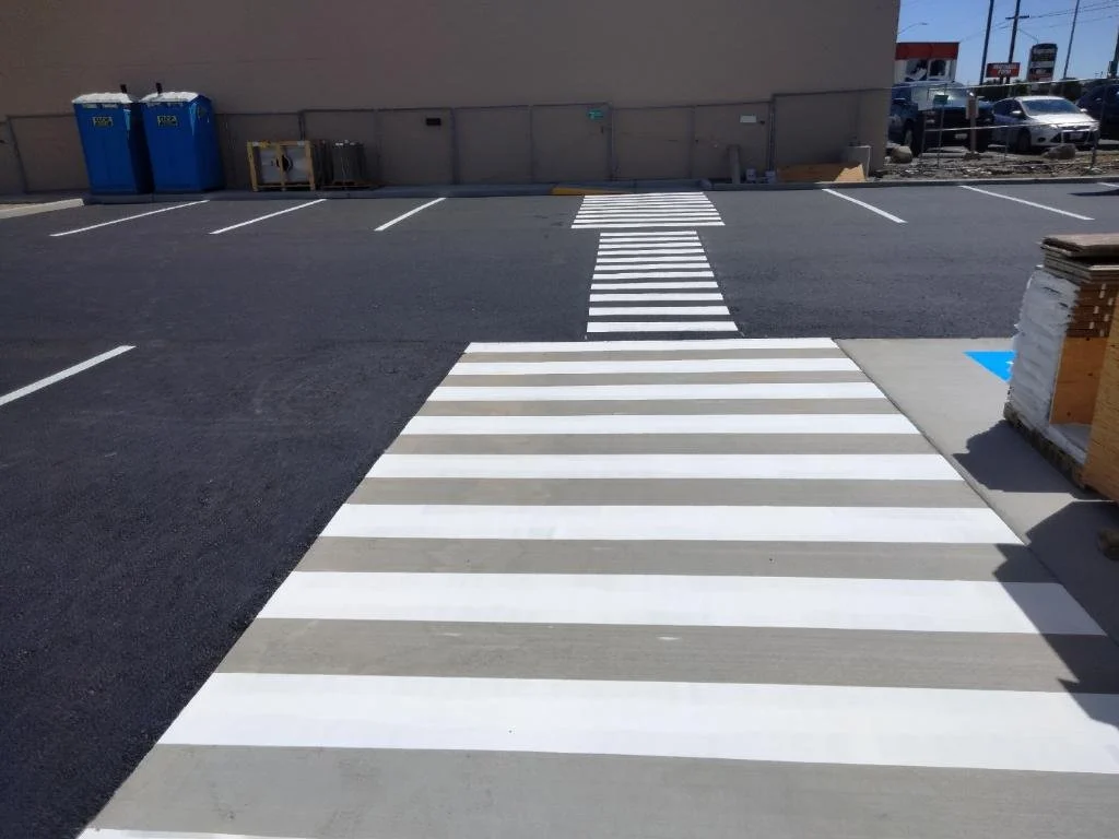 Pedestrian crosswalk with white stripes painted on the pavement in a parking lot, leading toward a building with a wall and vending machines in the background.