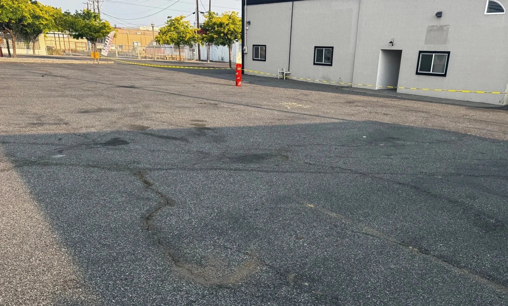 Empty parking lot with cracked asphalt, a gray building with several windows and a doorway, yellow caution tape along the edge, and a few trees in the background.