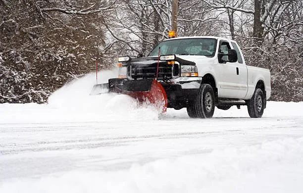 A white snowplow truck clearing snow on a road with snow-covered trees in the background.