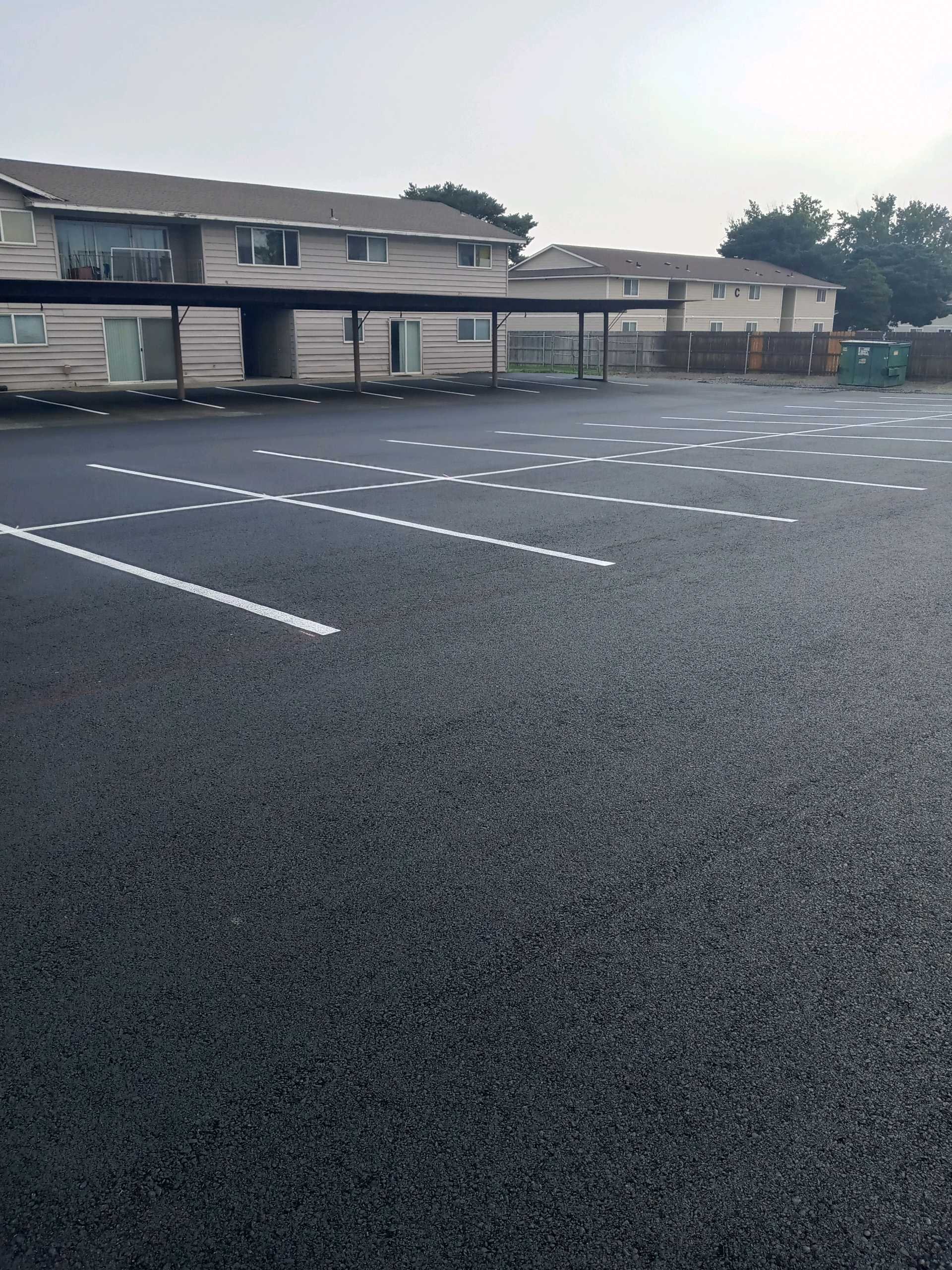 Empty parking lot with white painted lines, residential buildings in the background, and a cloudy sky.