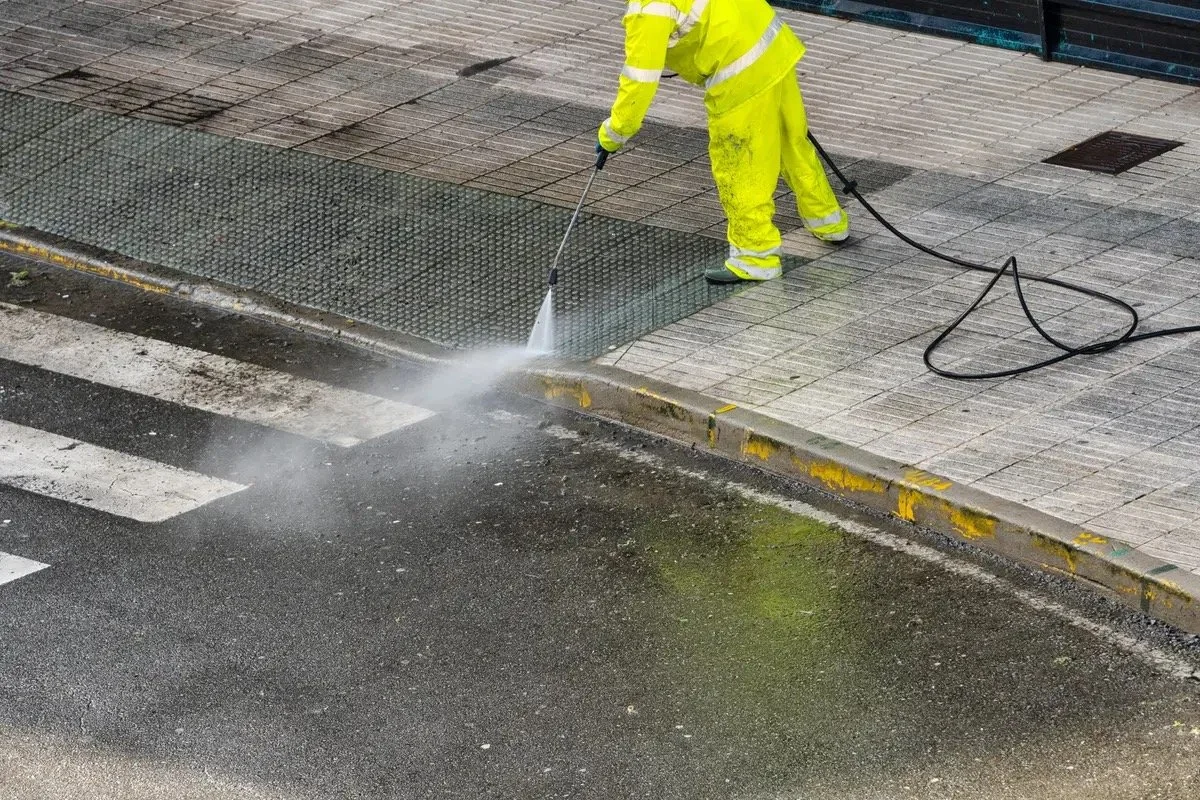 Worker in yellow safety gear pressure washing a sidewalk near a crosswalk on a city street.