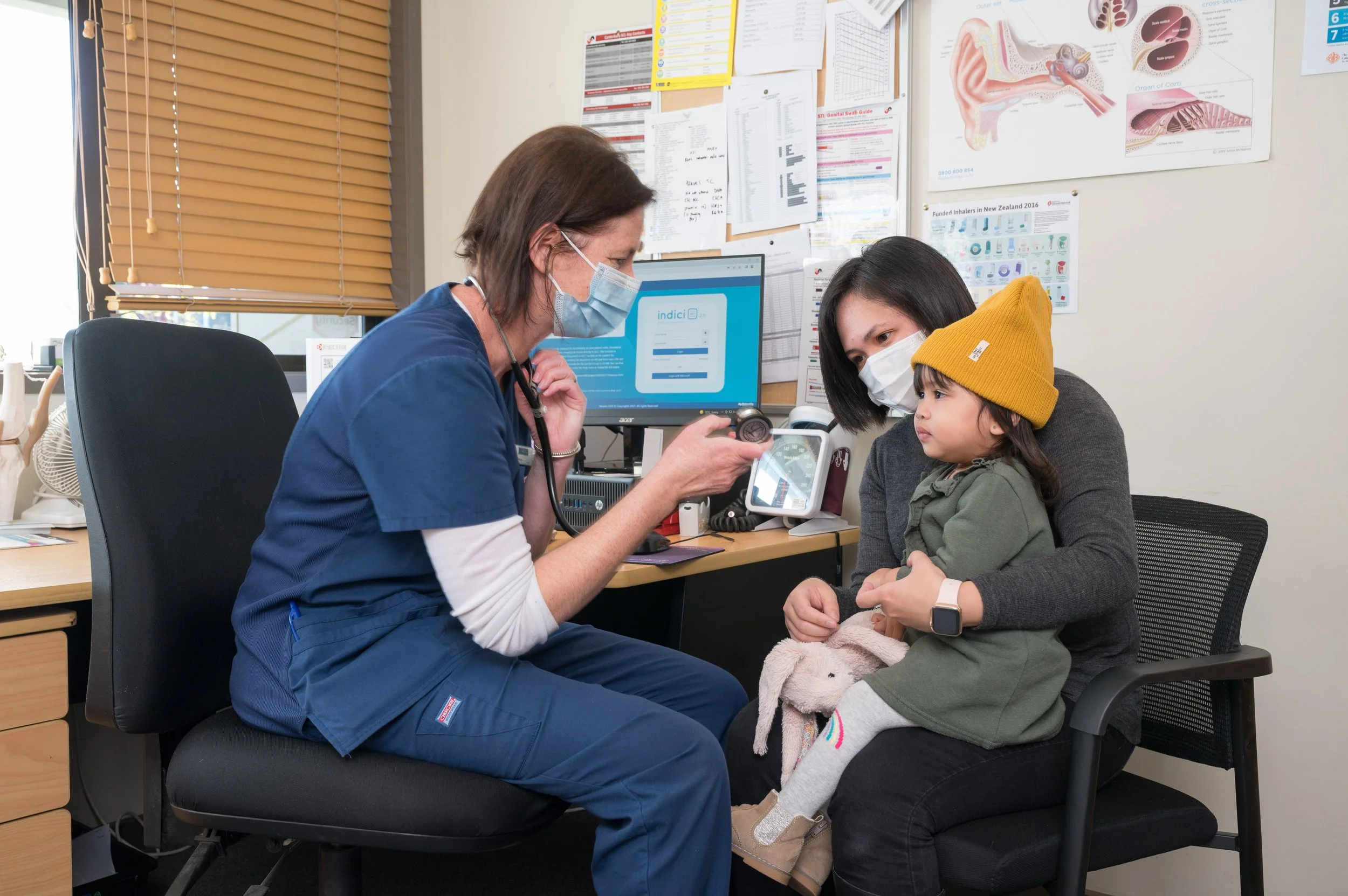 A healthcare professional, wearing a blue uniform and a face mask, examining a young girl with a stethoscope in a medical office. The girl, sitting with her mother, is wearing a yellow hat and holding a plush toy, while the mother is wearing a gray sweater and mask.