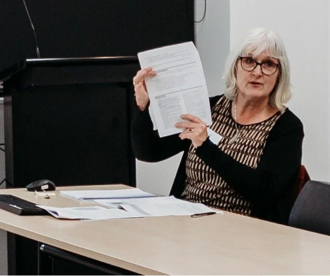 Woman with gray hair and glasses sitting at a table, holding up a document, in a classroom or office setting.