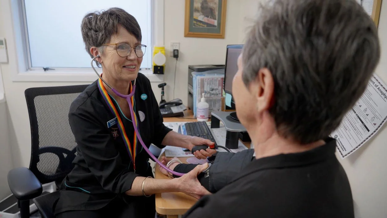 A healthcare professional with short gray hair, wearing glasses and a stethoscope around her neck, checks a patient’s blood pressure in an office. The professional is smiling and holding a blood pressure cuff while the patient has their arm extended. There is a computer, hand sanitizer, and framed pictures in the background.