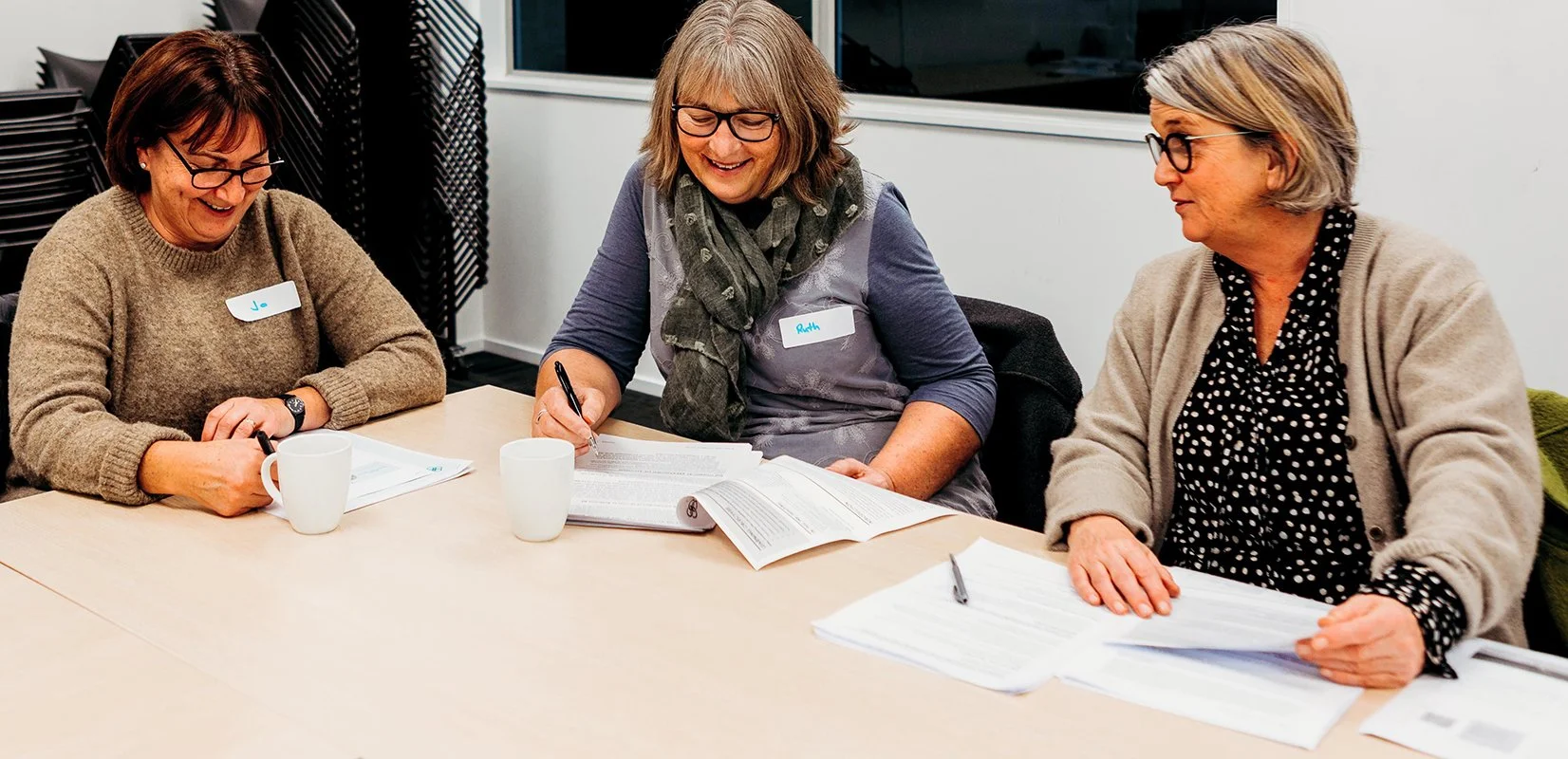Three middle-aged women sitting at a conference table smiling and talking during a meeting. They have papers, notebooks, and cups in front of them.