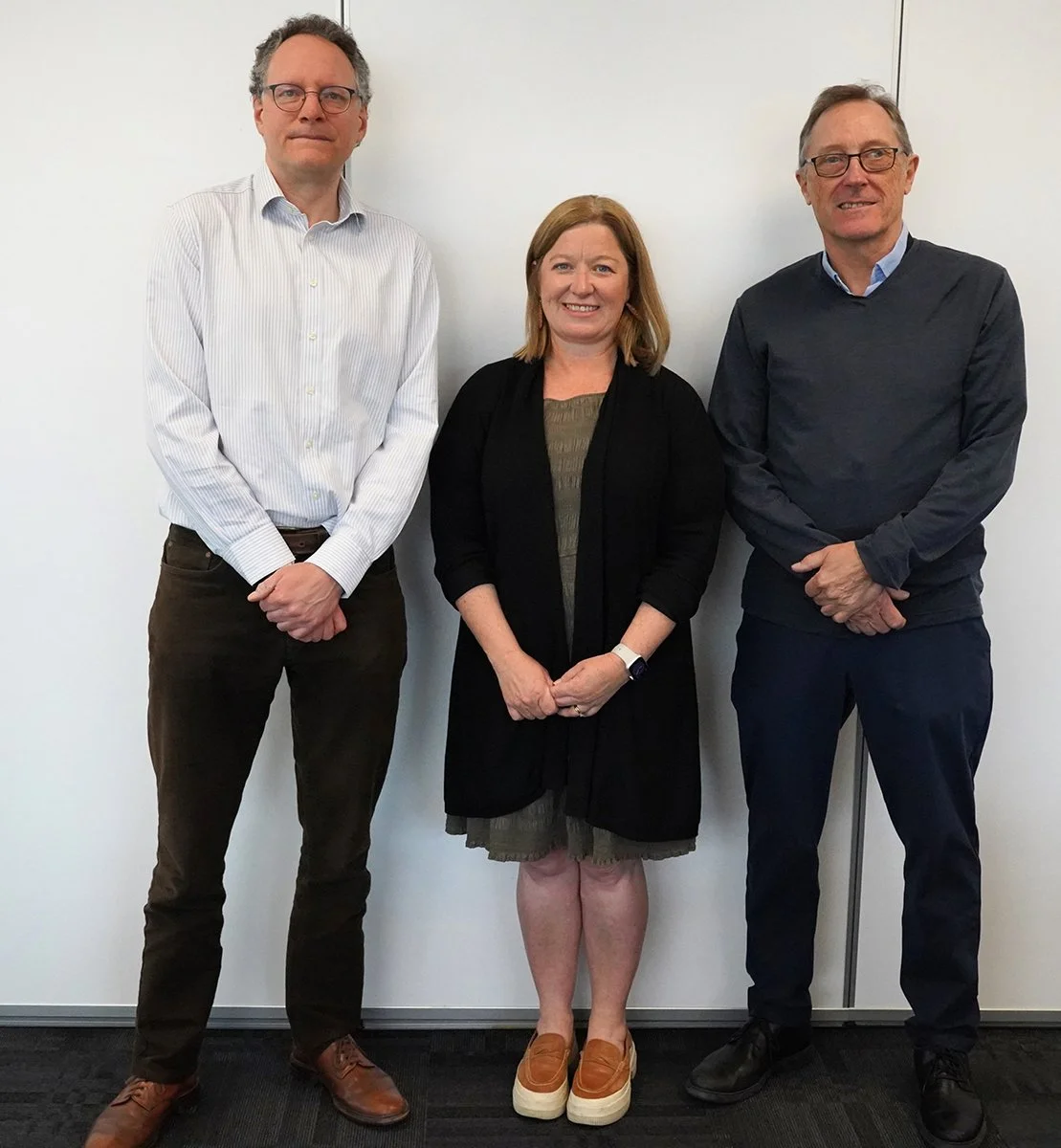 Three adults standing in front of a white wall, two men and one woman, dressed in business casual attire, smiling.