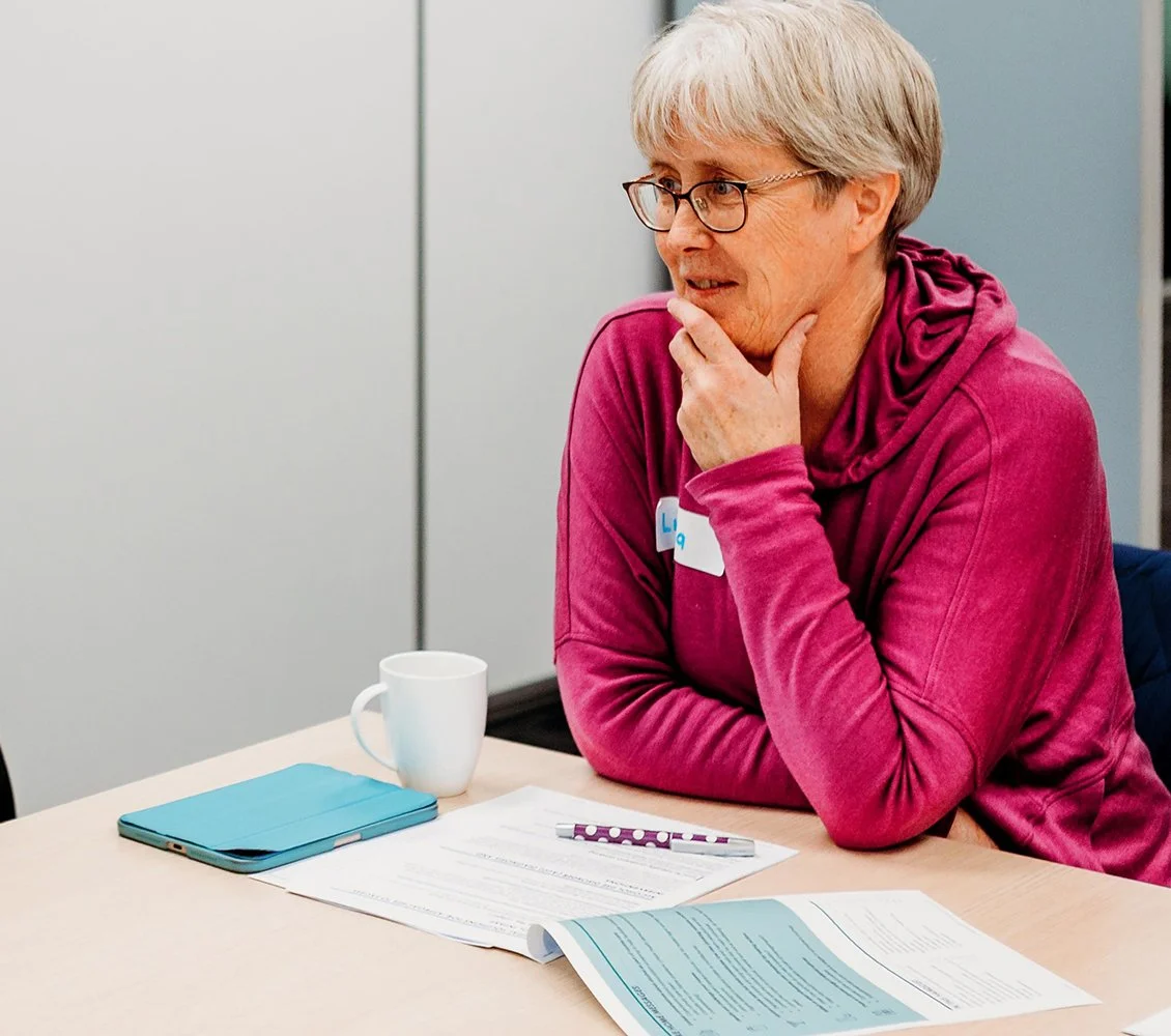 An older woman with short gray hair and glasses, wearing a pink hoodie, sits at a table with paperwork, a pen, a blue notebook, a white mug, and a name badge.