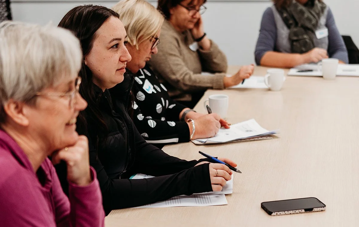 Group of women sitting at a conference table, taking notes during a meeting with notebooks, pens, mugs, and a phone on the table.