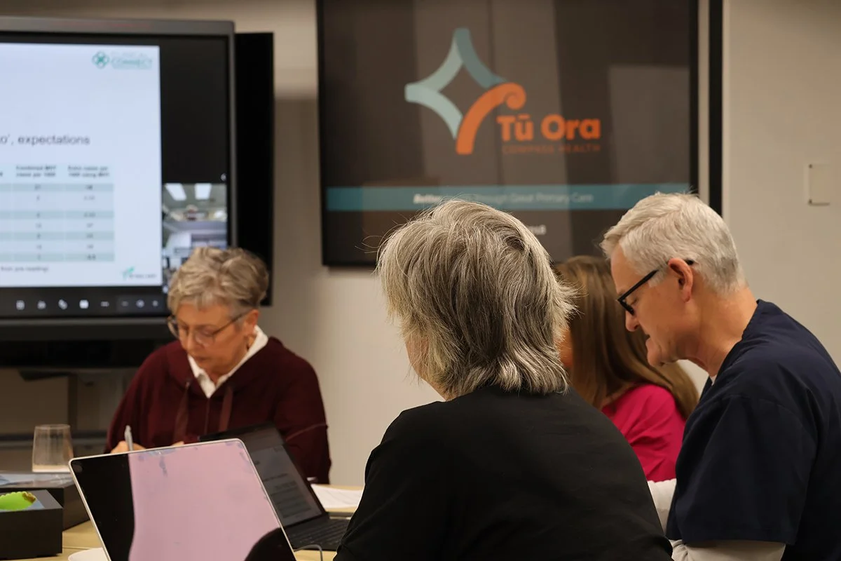 Group of middle-aged people in a meeting room, working on laptops and taking notes, with a presentation screen and logo behind them.