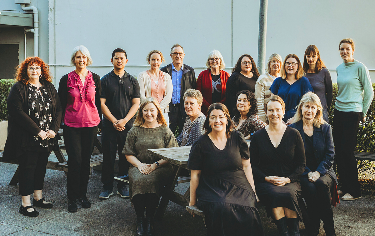 Group of diverse people, mostly women, gathered outdoors for a photo, standing and sitting around a picnic table.