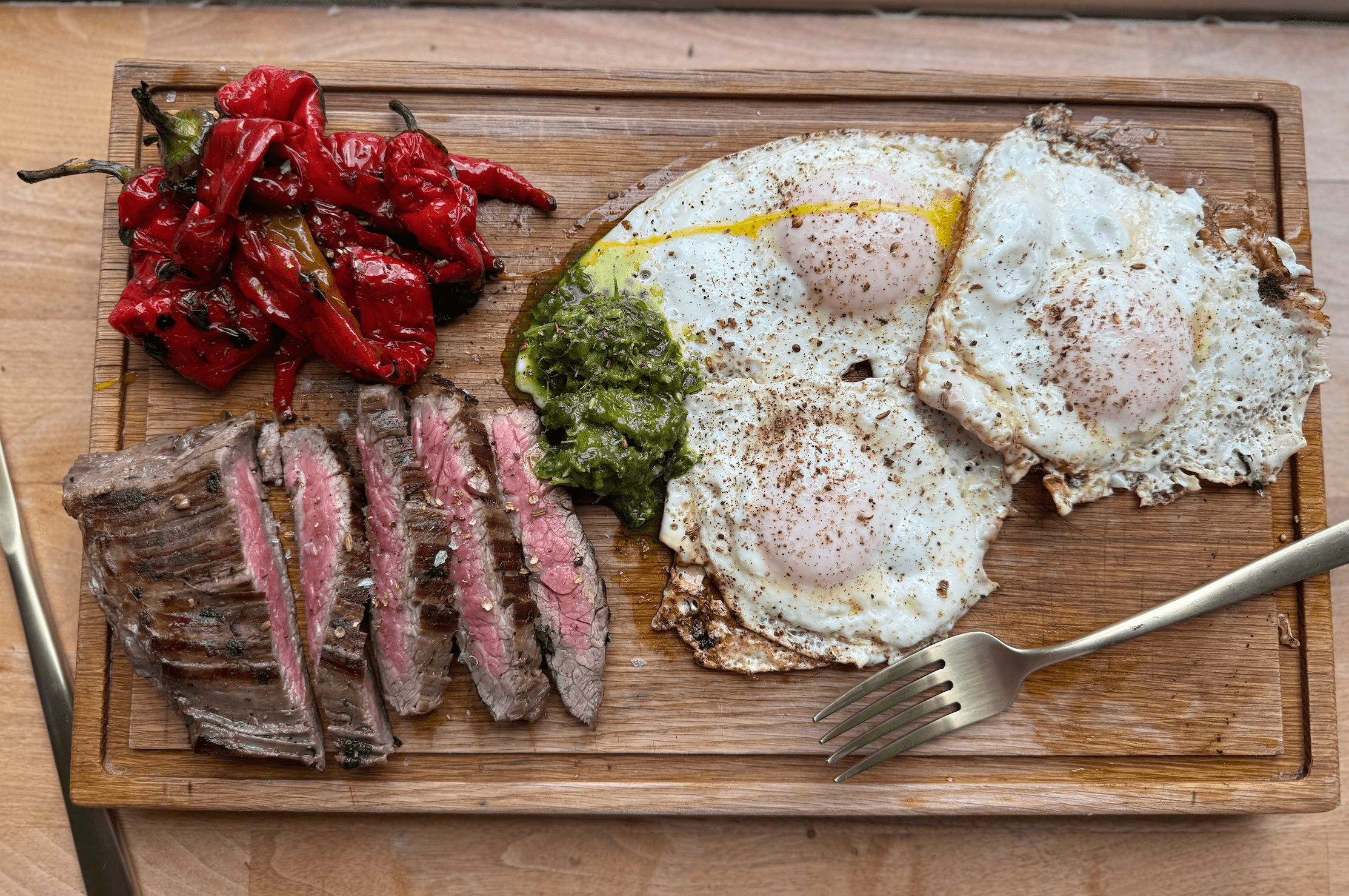 Wooden serving board with grilled steak slices, sunny side up eggs, roasted red peppers, and a dollop of green sauce.