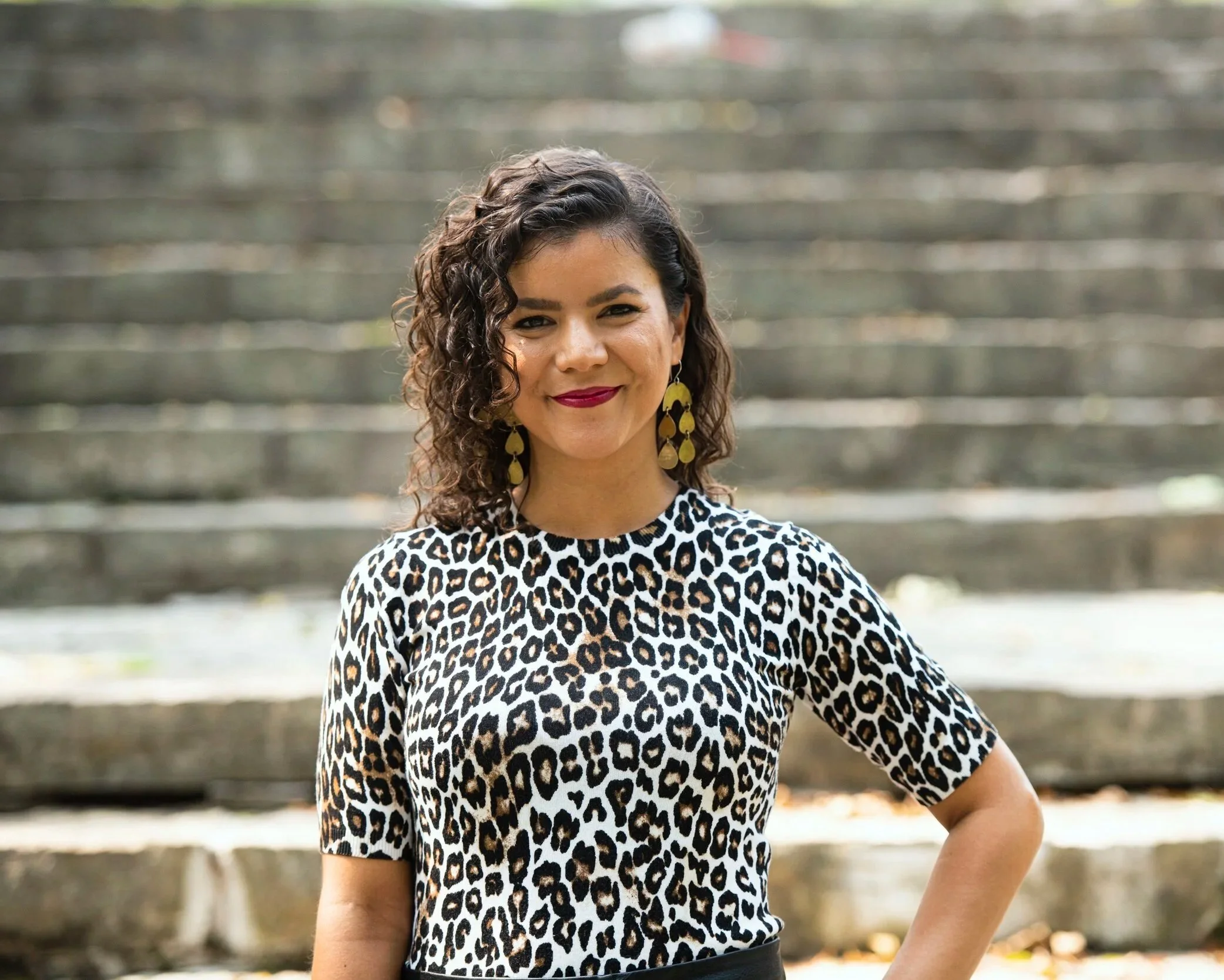 A woman with curly dark hair, wearing a leopard print top, gold earrings, and red lipstick, standing outdoors on stone steps and smiling at the camera.