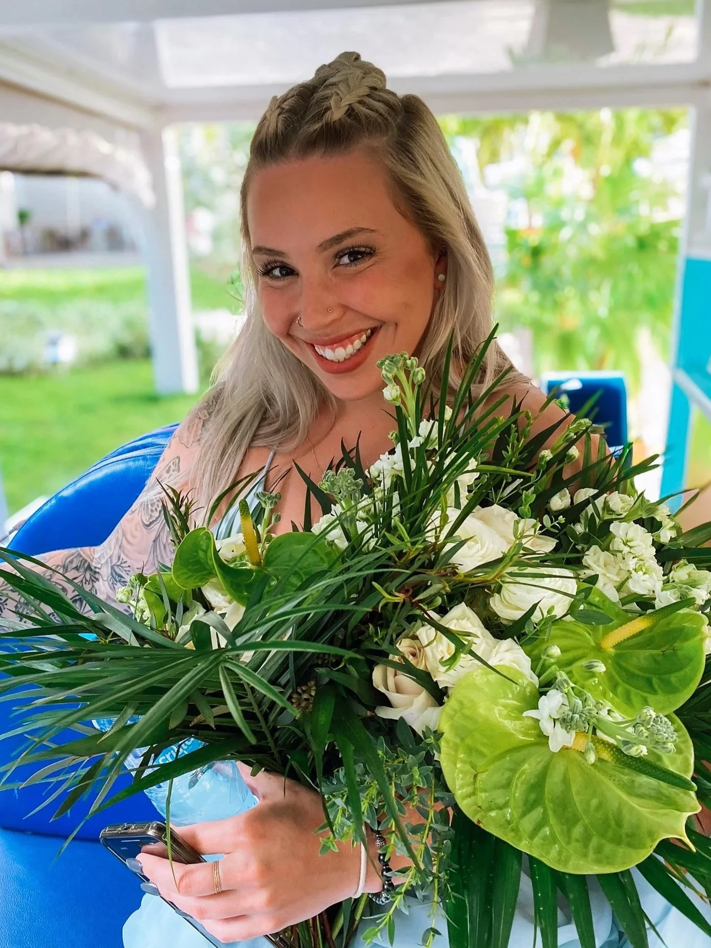 A young woman with blonde hair, smiling, holding a large bouquet of white flowers and greenery, sitting outside on a sunny day.