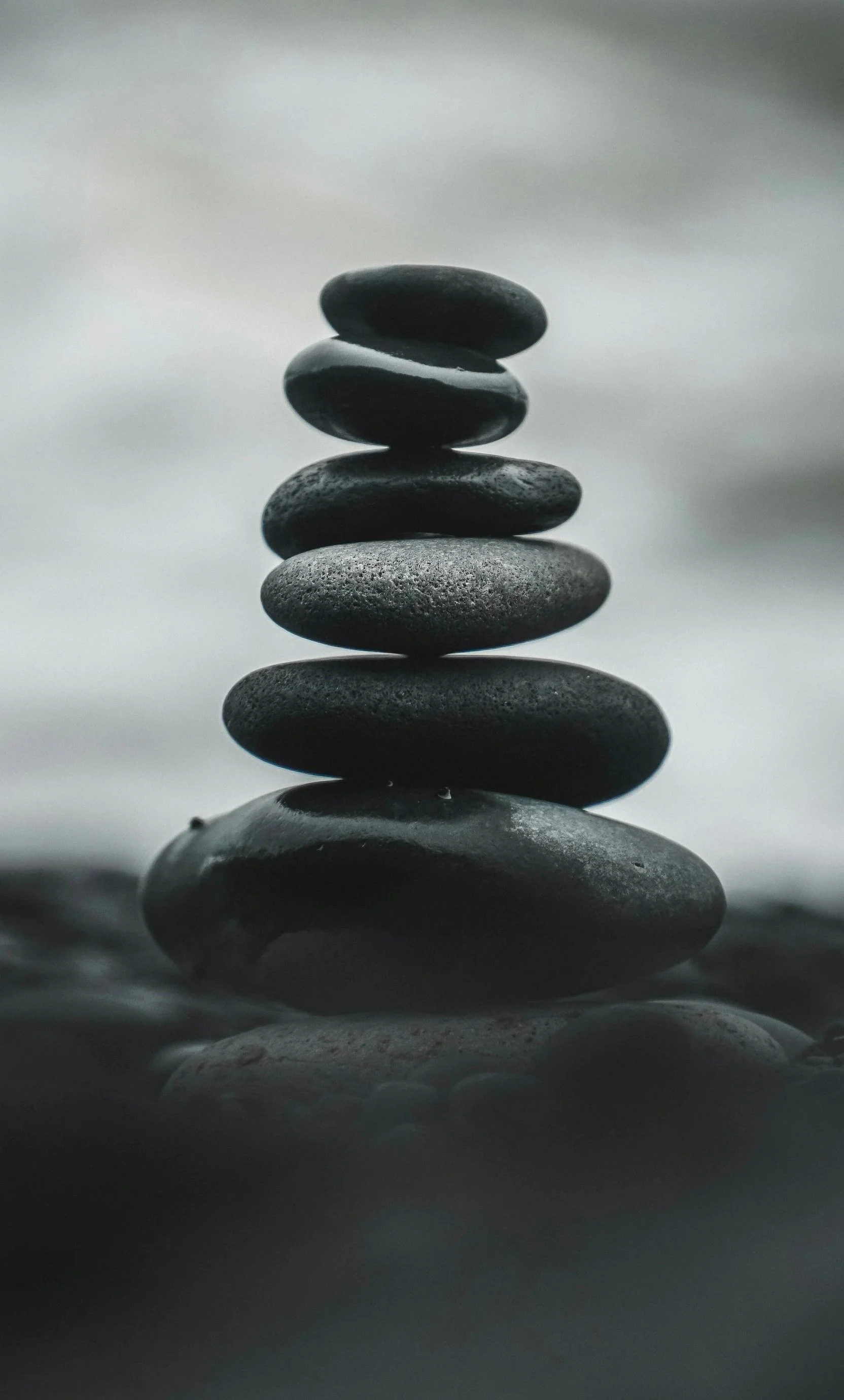 A stack of seven smooth, dark stones balanced on top of each other, set against a blurred, cloudy gray sky background.