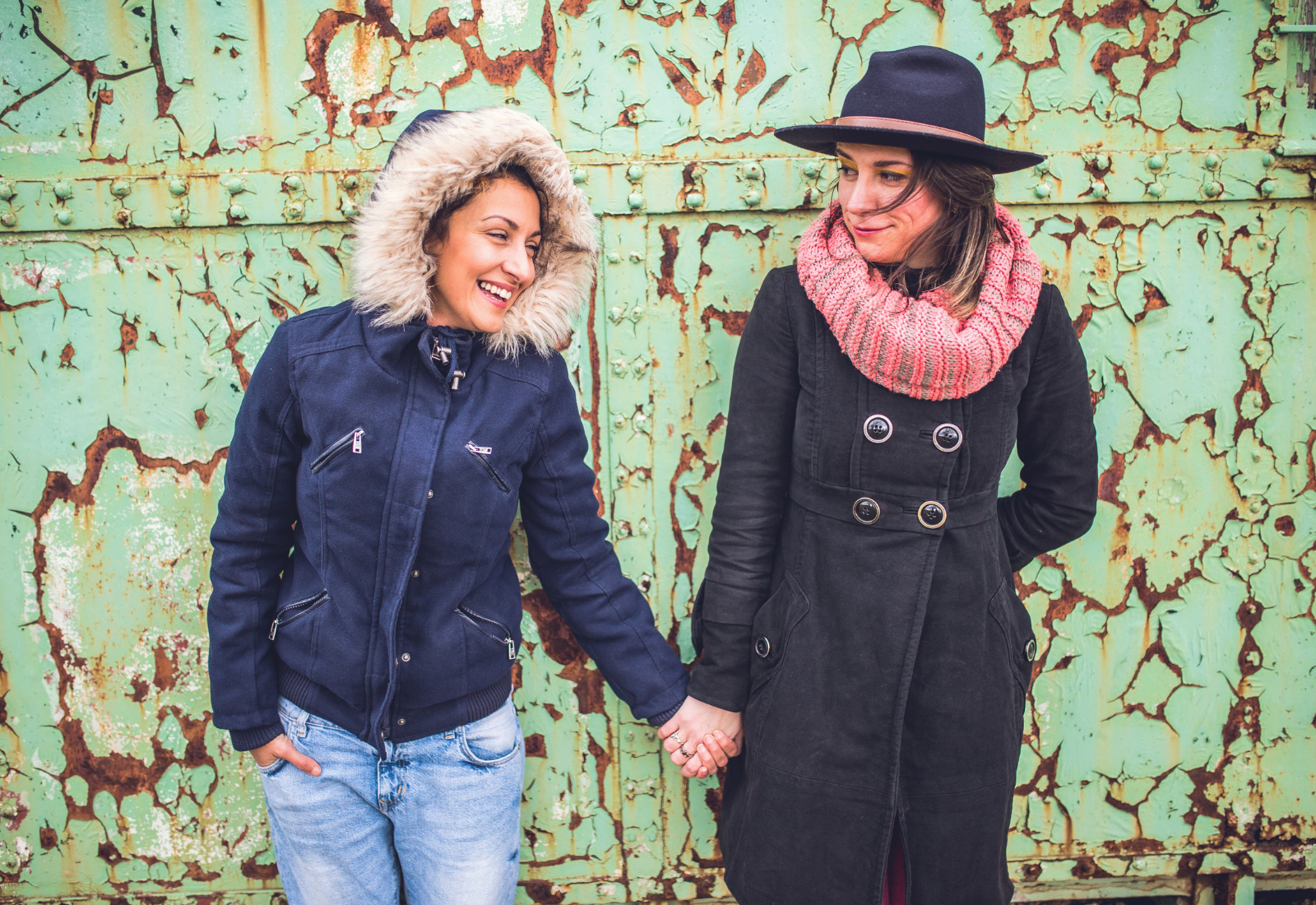 Two women standing and holding hands in front of a rusted, green metal wall. The woman on the left is smiling, wearing a navy jacket with fur hood, and light jeans. The woman on the right has a shy smile, wearing a black coat with large buttons, a red and pink scarf, and a black hat.