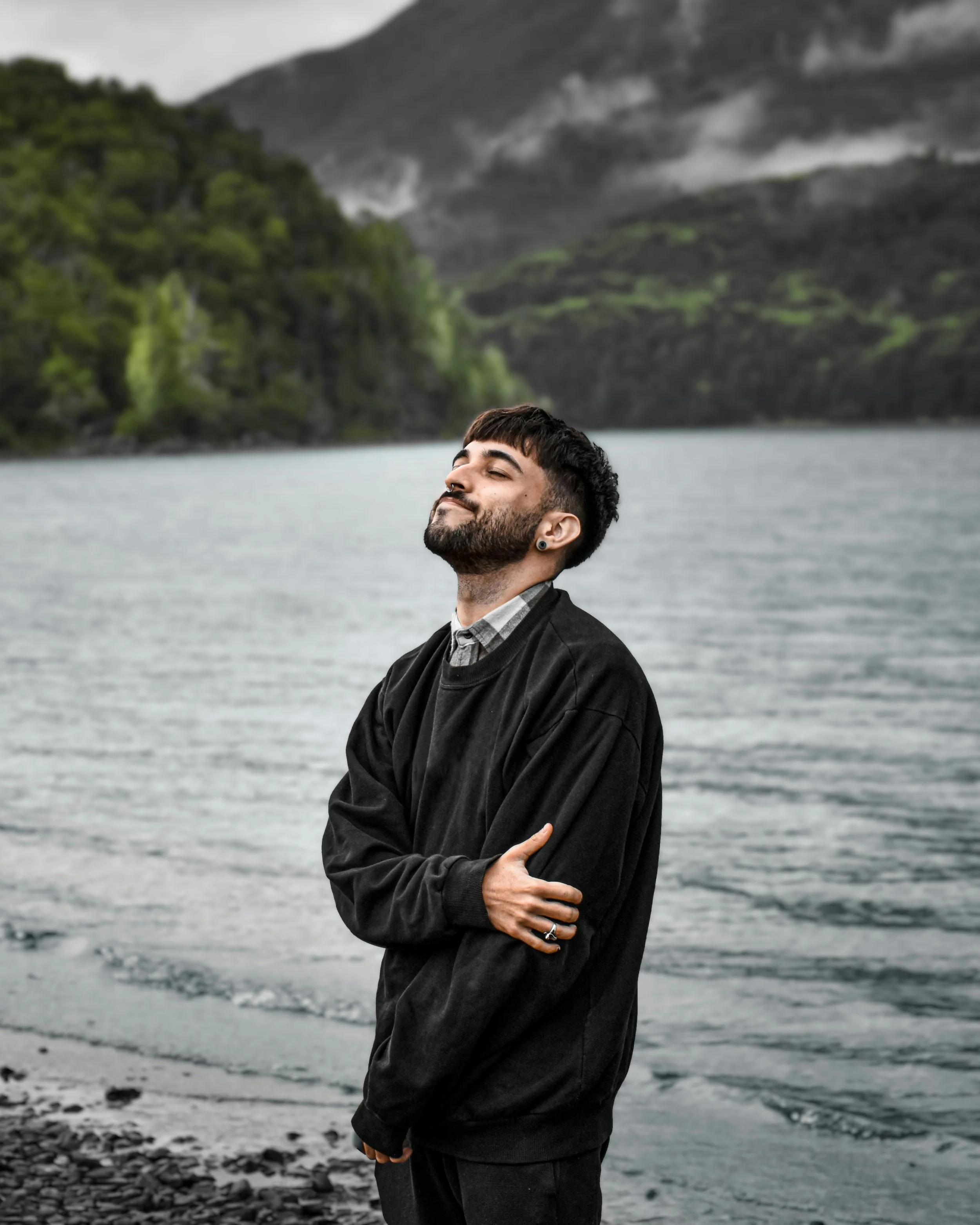 A young man with a beard and earrings standing by a lake with mountains in the background, smiling with eyes closed, arms crossed, wearing a black sweatshirt and a checked shirt.