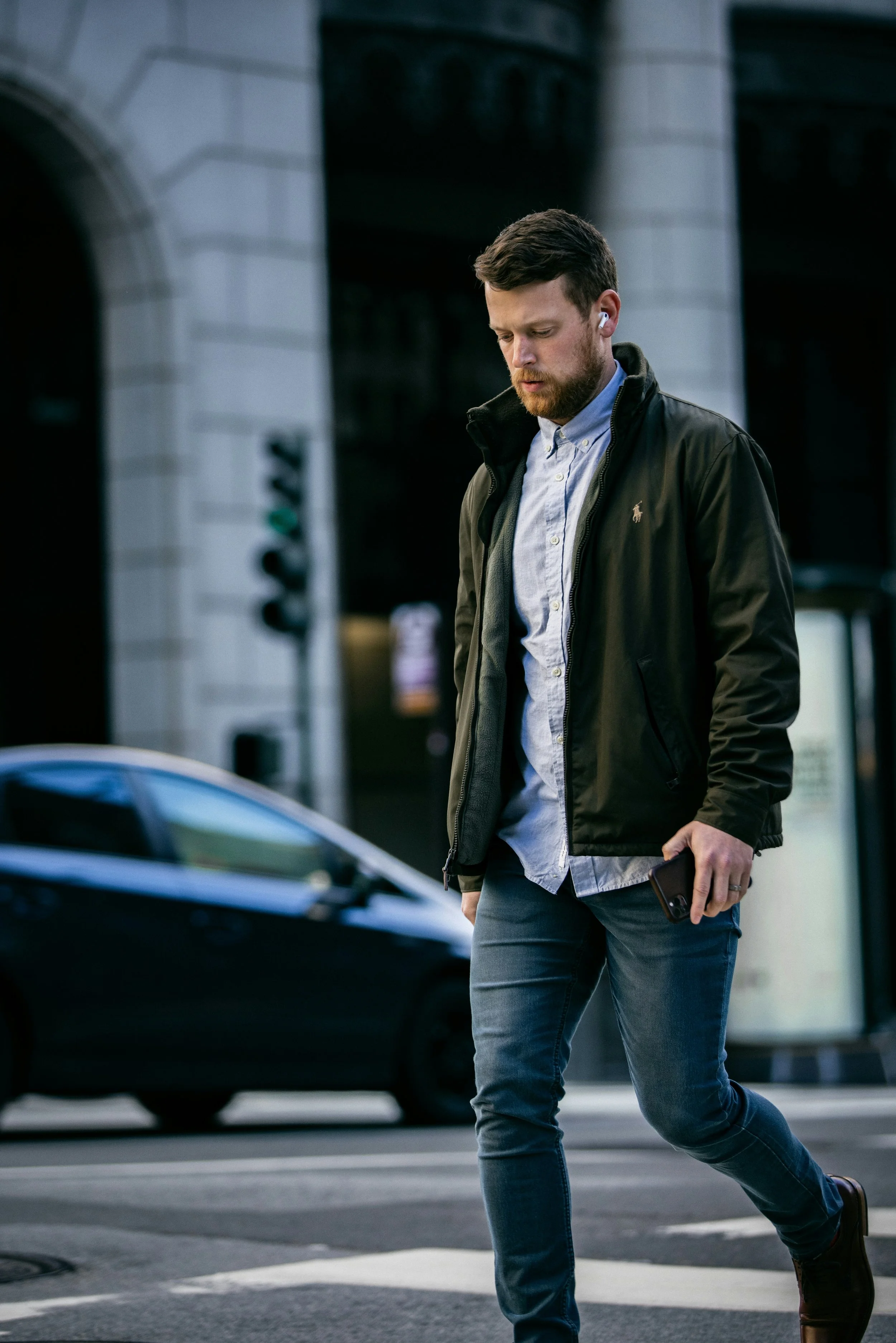 A man with a beard and short brown hair, wearing a black jacket, light blue button-up shirt, and jeans, walking across a city street while holding a smartphone in his right hand.