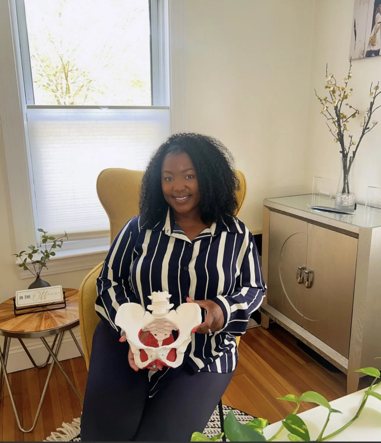 A woman sitting on a yellow chair in a room, holding a model of a pelvis. She is smiling and wearing a navy and white striped shirt. There is a side table with a small plant and a sign that reads 'In the Office' on it, a cabinet with decorations, and a window with natural light.