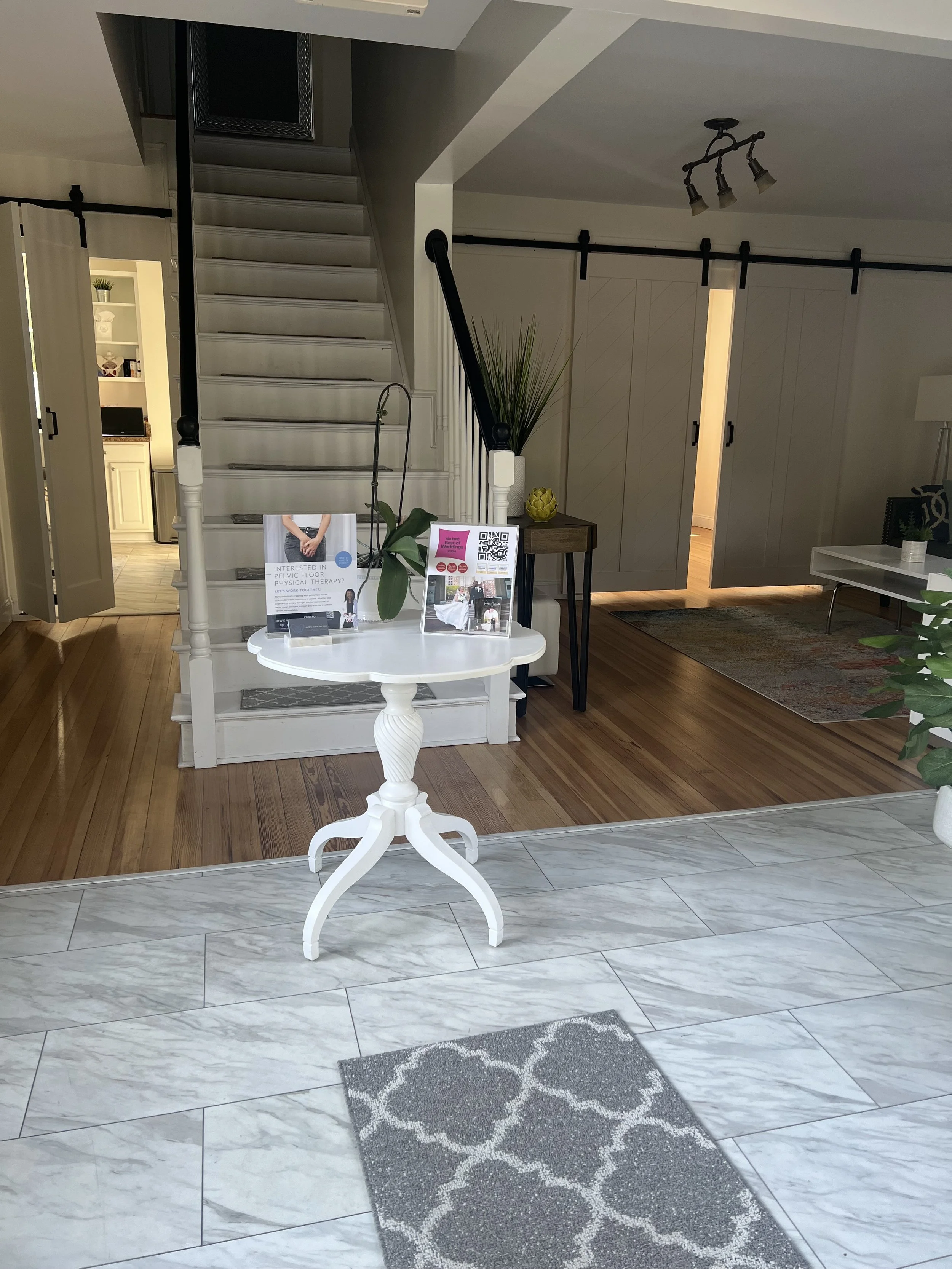 Indoor view of a home entryway with a small round white table holding brochures and a plant, a staircase with white railings, and a wooden floor leading to a hallway with sliding barn doors, a white cabinet, and a rug.