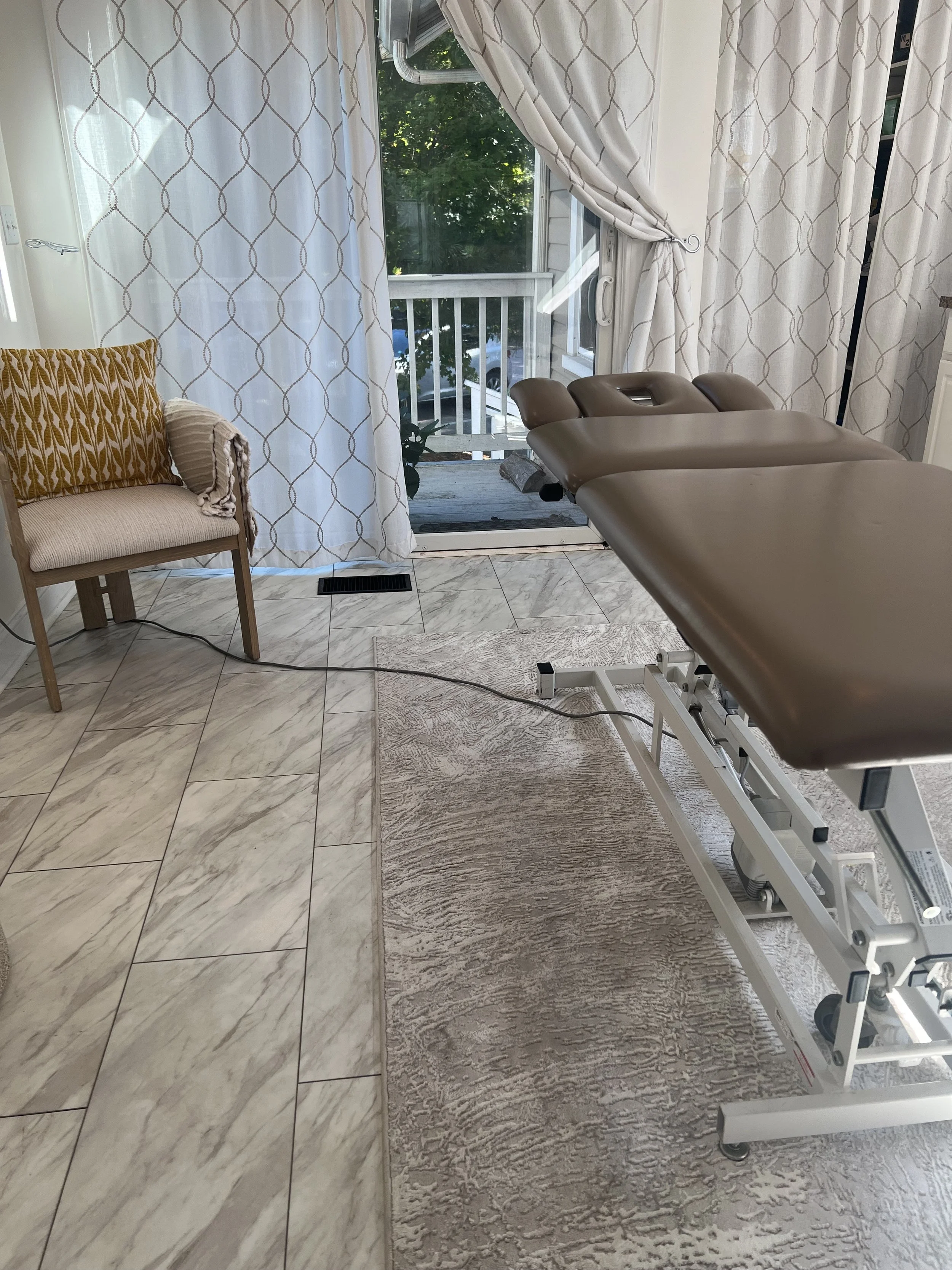 A medical examination room with a brown examination table, a beige armchair with a yellow pillow, white curtains with a geometric pattern, a sliding glass door leading outside, and a patterned rug on a marble floor. Pelvic Floor Physical Therapy trea