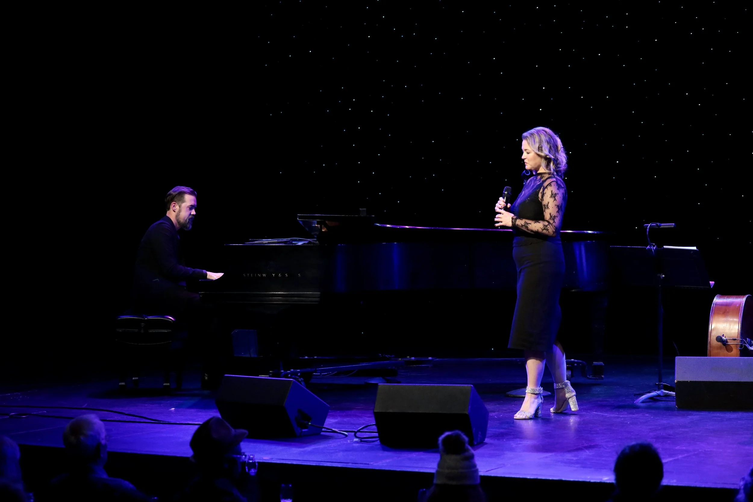 A woman in a black dress with lace sleeves and high heels performs on stage, singing into a microphone while a man in a black suit plays a grand piano beside her. The stage has black curtains with small white lights, and there is audience watching.
