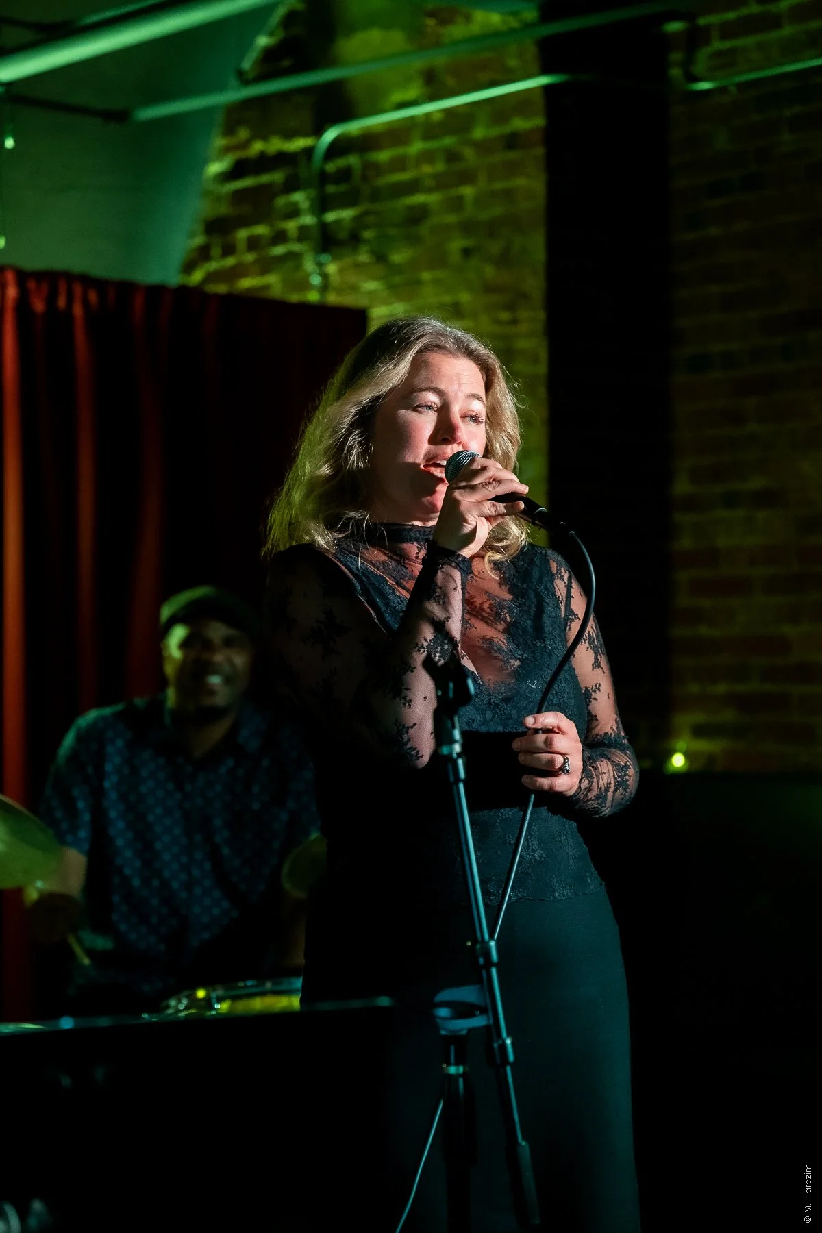 Stina Tillotson, Seattle Jazz Vocalist. A woman singing into a microphone on stage with a man playing drums in the background, in a dimly lit venue with brick walls and green lighting.
