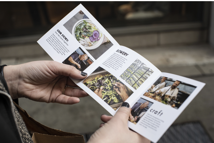 Person holding a folded brochure or pamphlet with images and text about gardening or farming, outdoors setting.