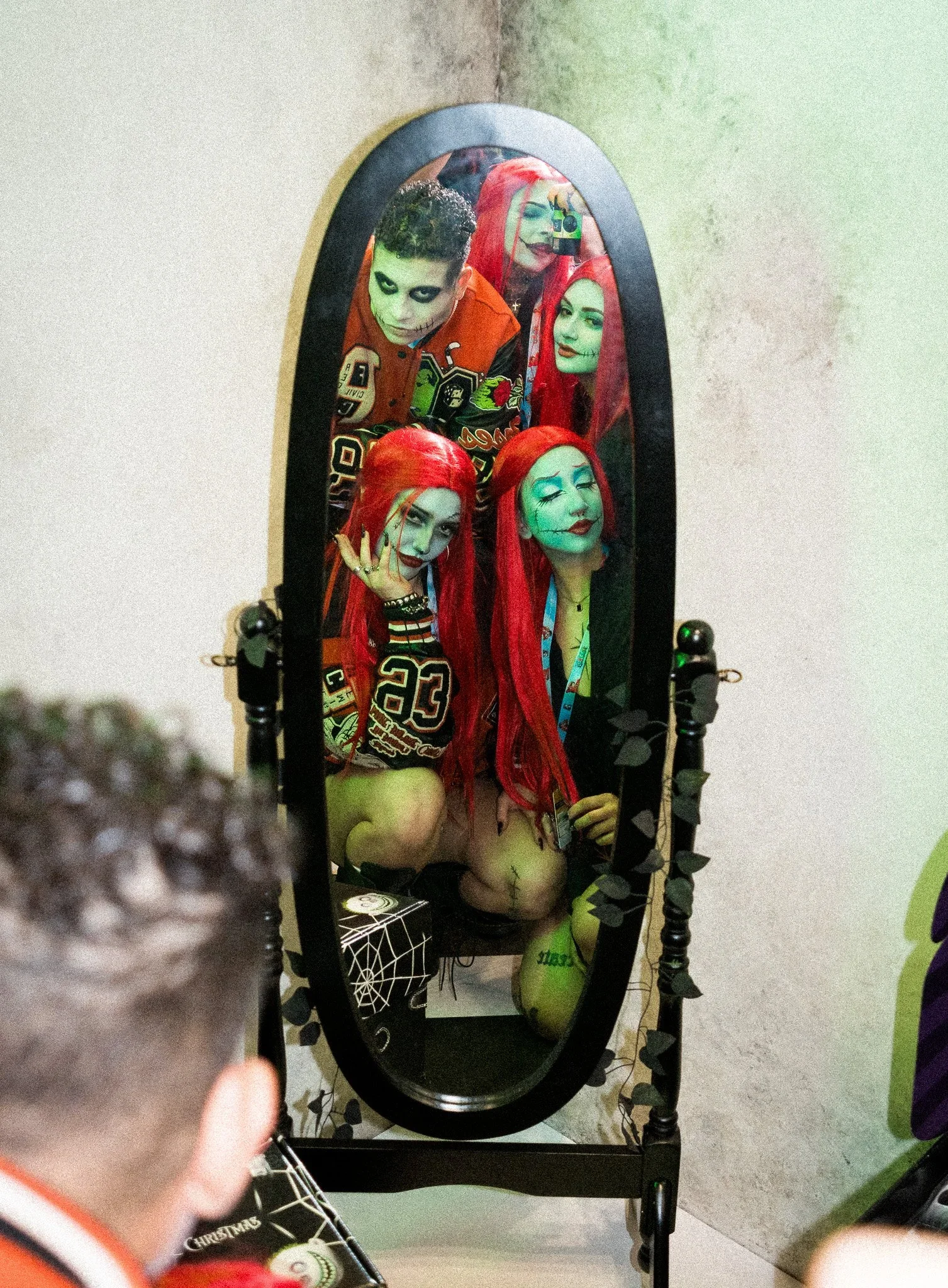 Person with face paint and Halloween costume taking a selfie in front of a mirror featuring other costume characters. New York Comic Con 2024 booth. Event production and marketing by Laura Alyse Rubin.
