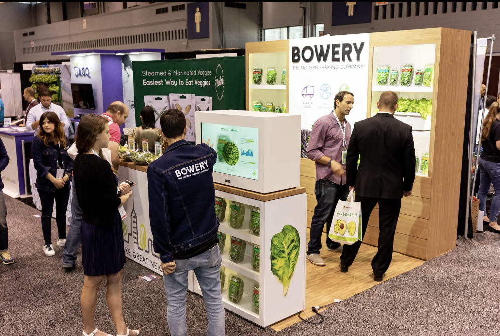 Trade show booth for Bowery, a modern farming company specializing in vegetables, with displays of packaged greens and conversation among attendees. Laura Alyse Rubin marketing and event producer.