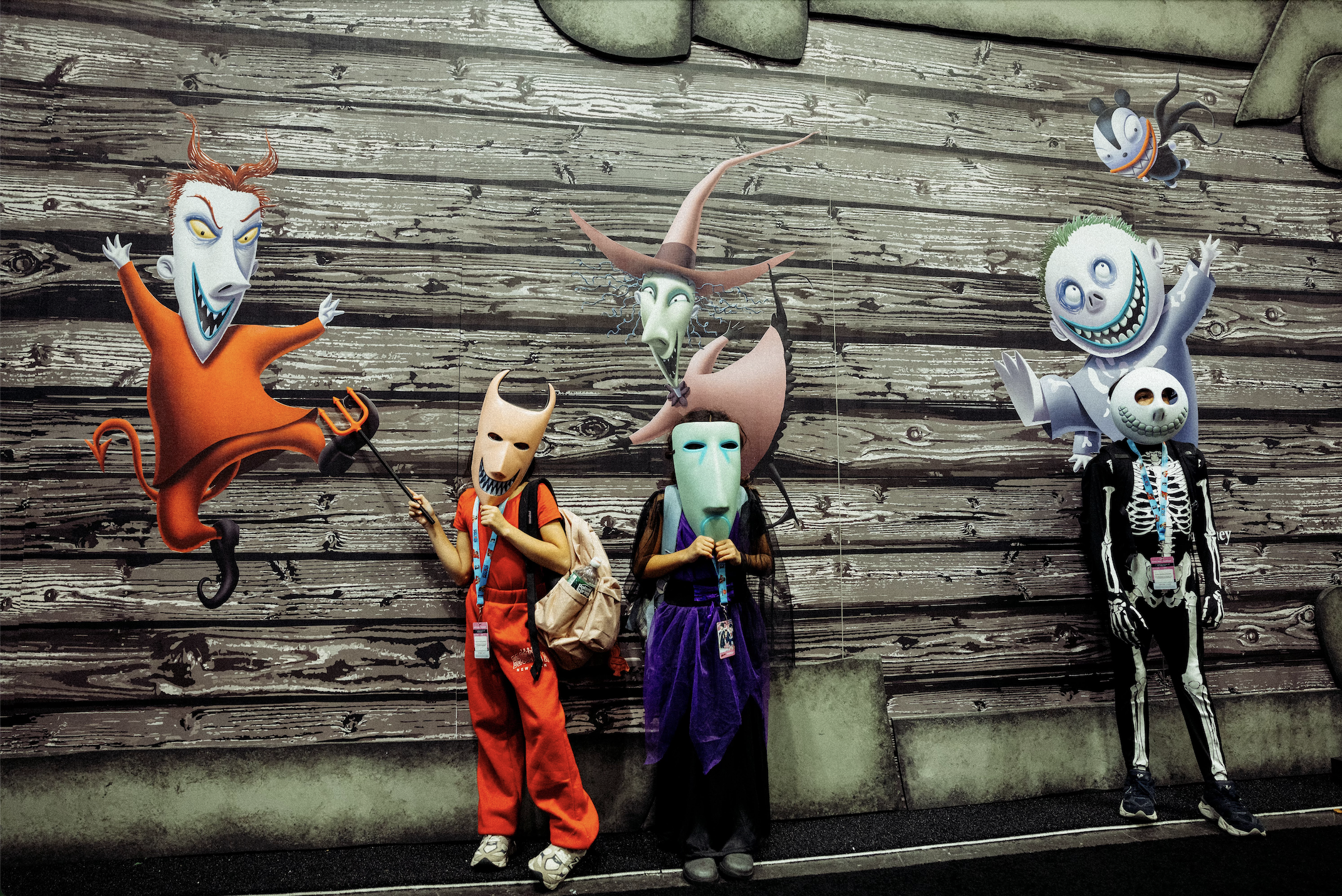 Three children wearing mascot masks featuring characters from Tim Burton's "The Nightmare Before Christmas" standing against the Civil Regime booth at New York Comic Con 2024. Event production and marketing by Laura Alyse Rubin.