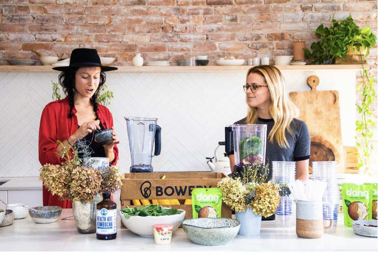 Rachelle Robinett in a modern kitchen with brick wall and potted plants, surrounded by bowls, bottles, and ingredients. Bowery Farming event activation produced by Laura Alyse Rubin along with Dang Chips and Health Aid Kombucha.