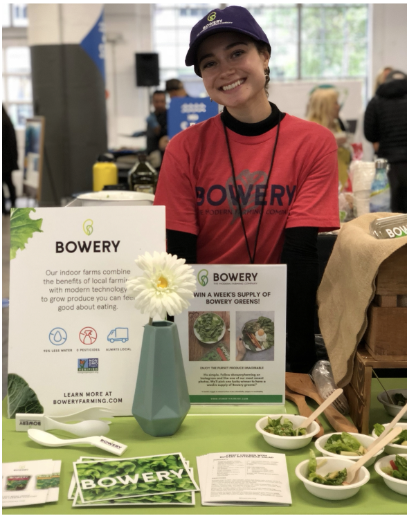 Smiling woman at Bowery Farming booth wearing red shirt and blue cap, surrounded by promotional materials and small bowls of greens at an indoor event.