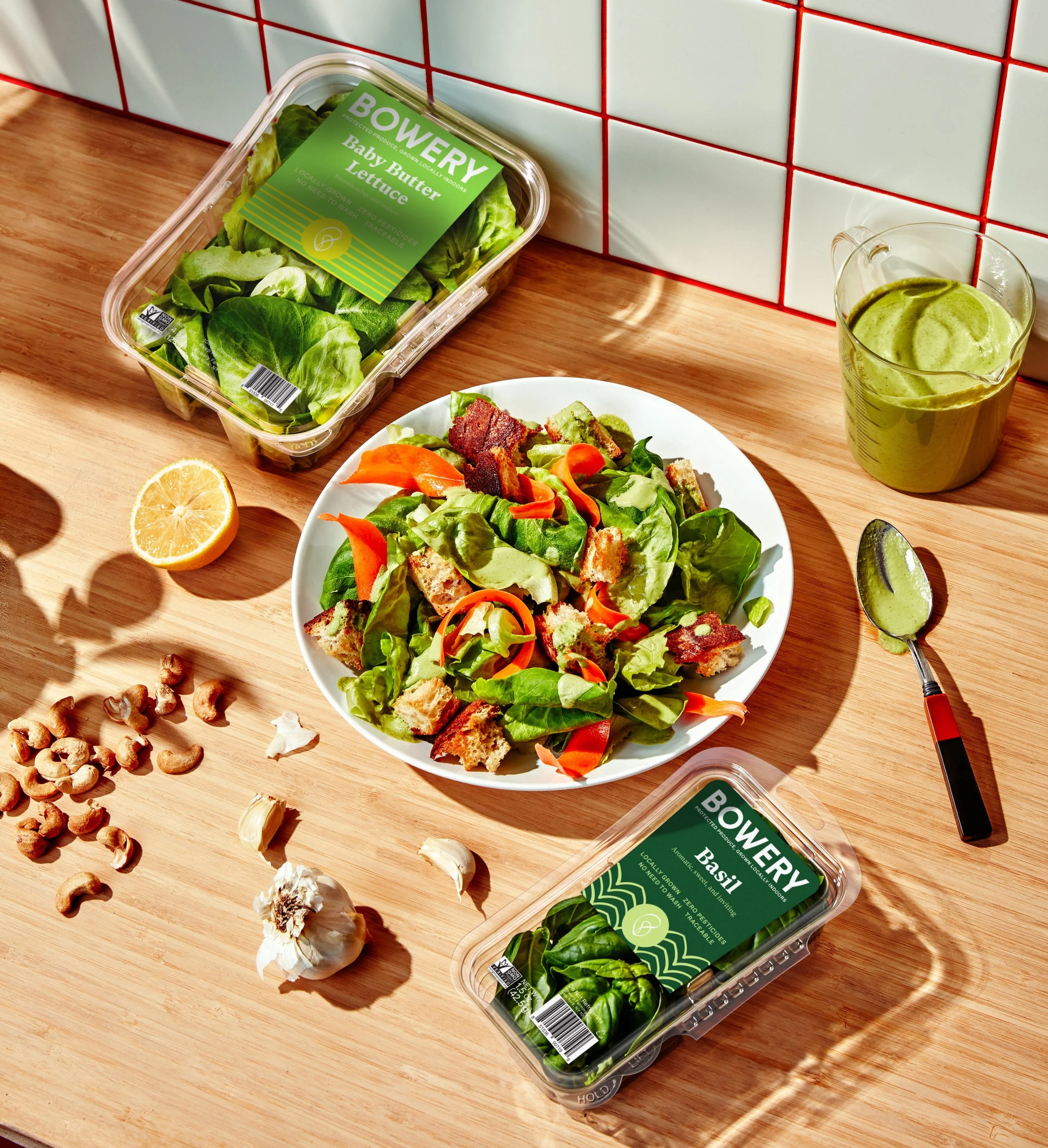 A wooden kitchen countertop with a salad in a white bowl, two containers of Bowery Farming hydroponic baby lettuce and basil, a halved lemon, a garlic bulb, some garlic cloves, and a few cashew nuts.
