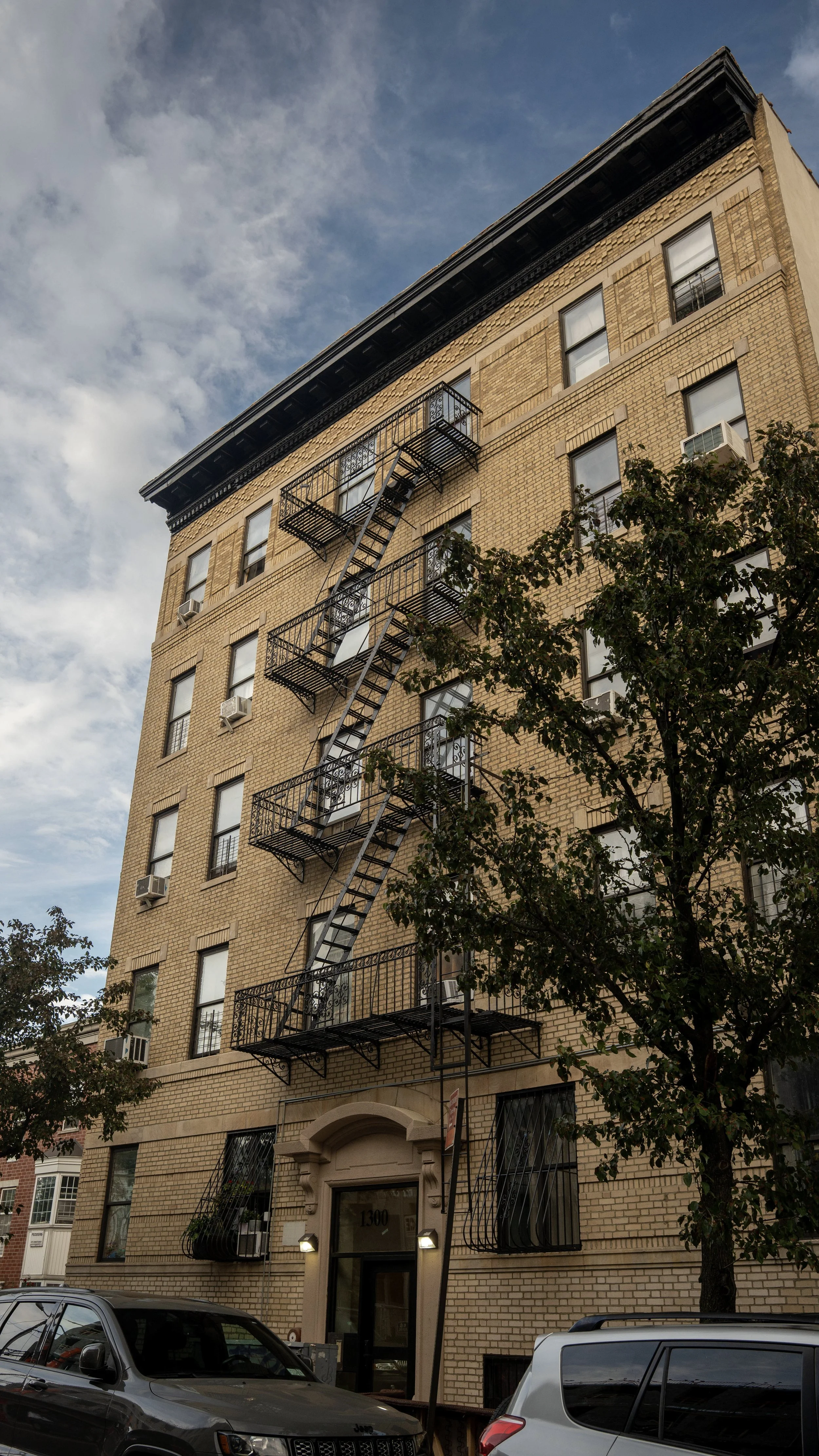 Six-story brick apartment building with fire escape stairs on the front, several windows with air conditioning units, and a decorative arched entrance at 1300 address. Trees and parked cars are in front.