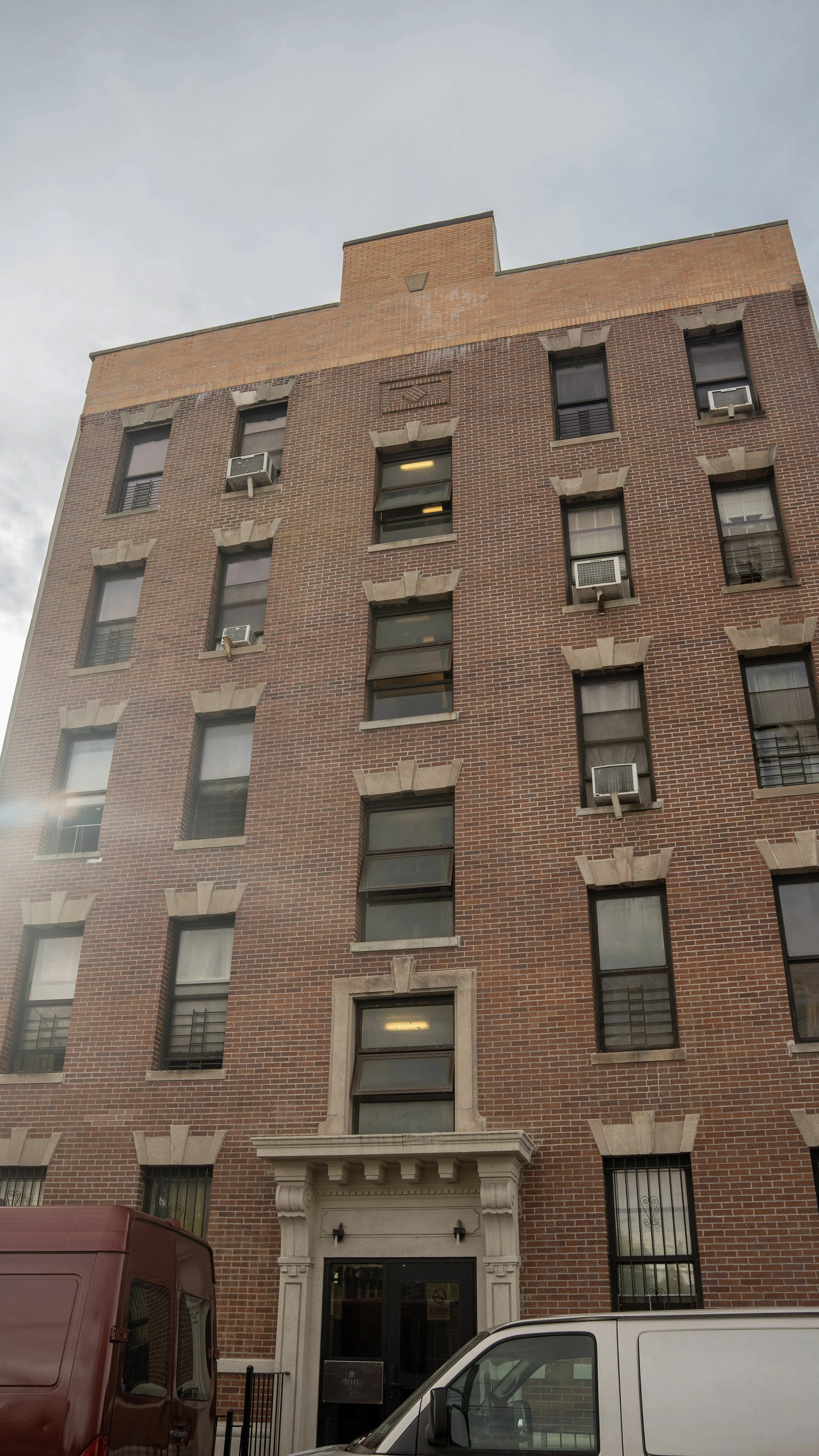 A tall red brick apartment building with multiple windows, some with air conditioning units, and a small entrance with an ornate stone frame. Vehicles are parked in front.