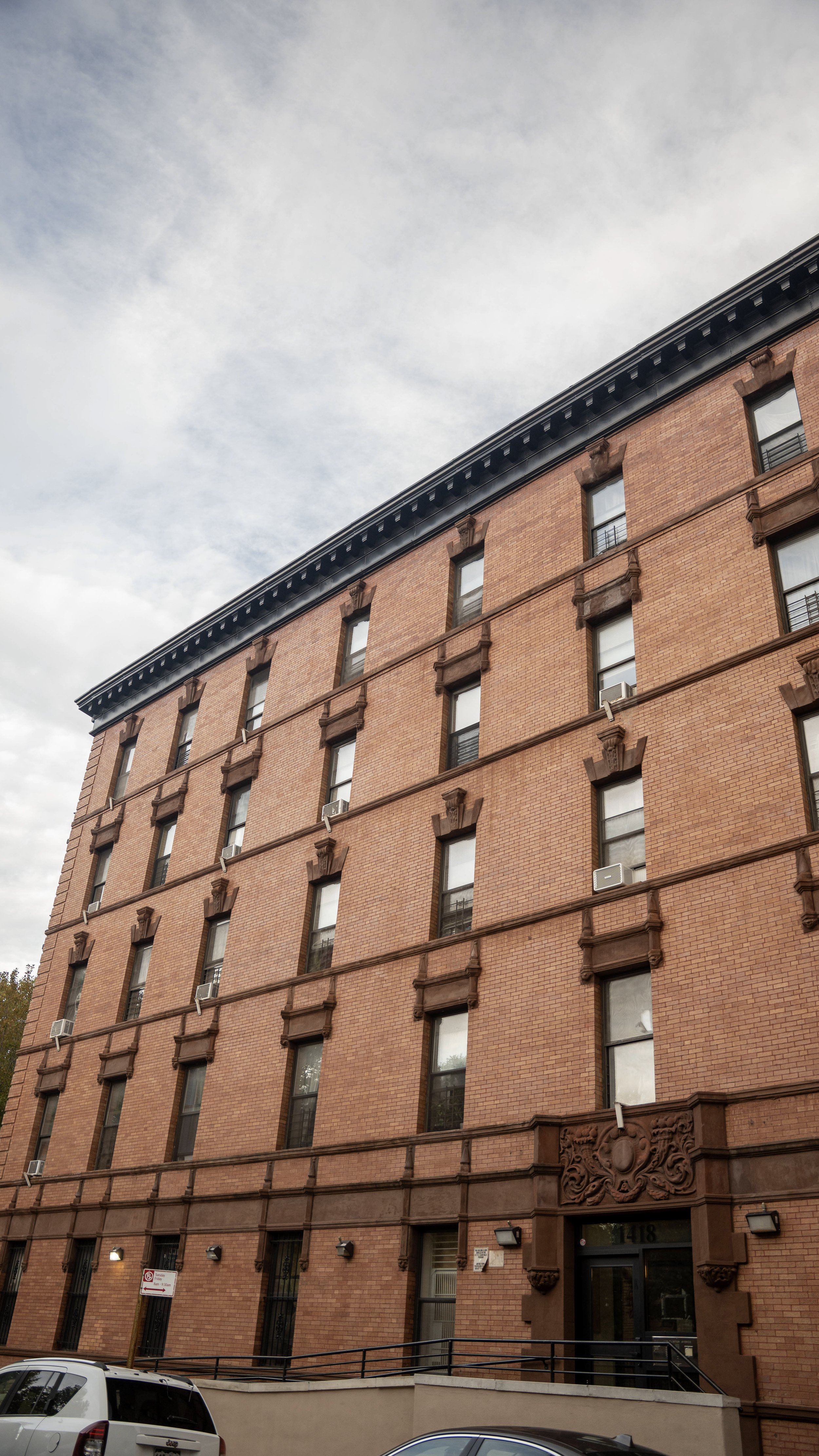 A multi-story brick building with decorative stone details above the windows and a black cornice on the roof, partially obscured by parked cars in the foreground and a cloudy sky above.