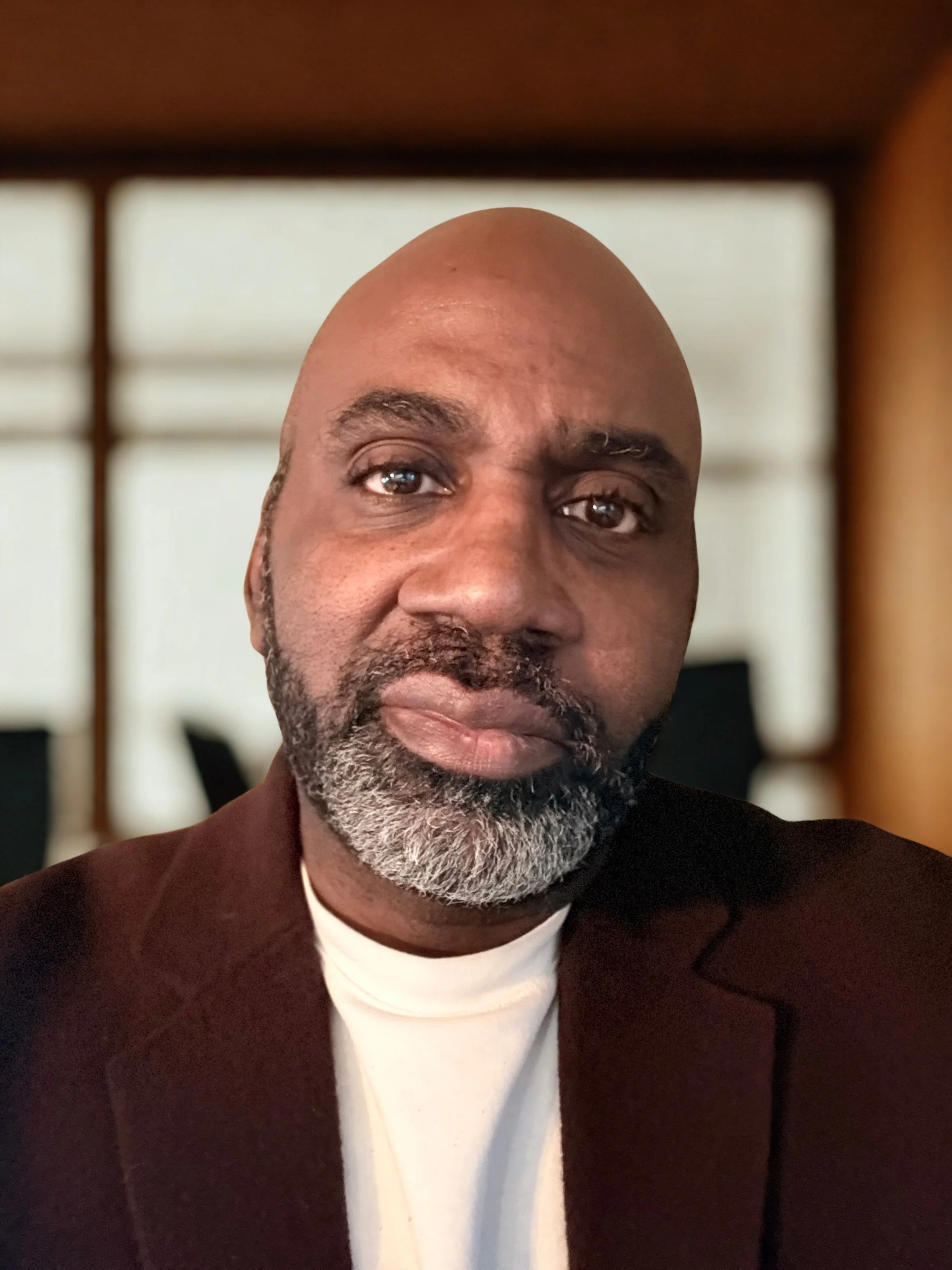 Close-up of a man's face with a bald head and gray beard, wearing a black jacket and white shirt, indoors with a blurred background.