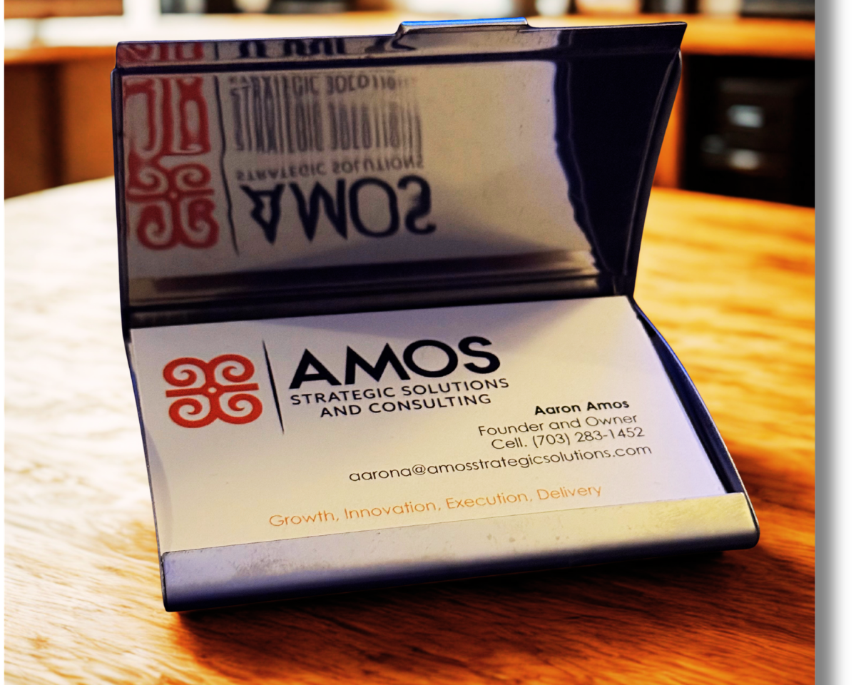 Open business card holder displaying a business card for Amos Strategic Solutions and Consulting, with contact details and services listed, on a wooden table.