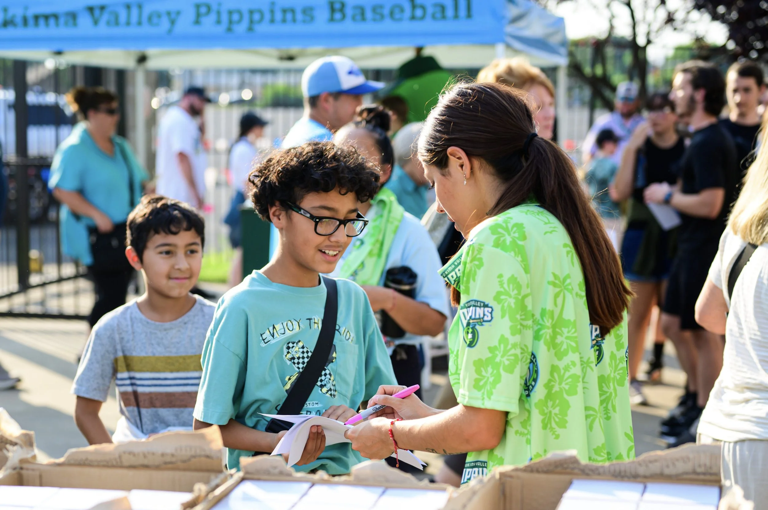A group of people at an outdoor event during daytime, with a banner reading 'Kimber Valley Pippins Baseball' visible in the background. Two children are interacting with a woman. The boy with glasses is holding a notebook, and the woman is signing it.