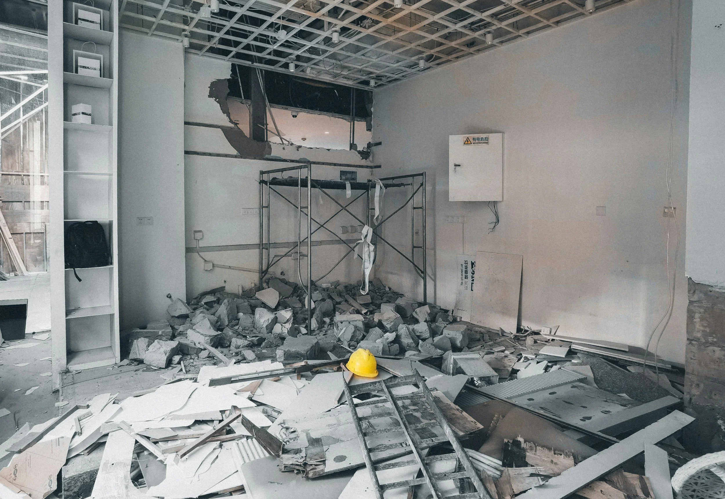 Interior of a building under construction or renovation with a large hole in the wall, scattered debris, a yellow safety helmet, a ladder, and construction scaffolding.