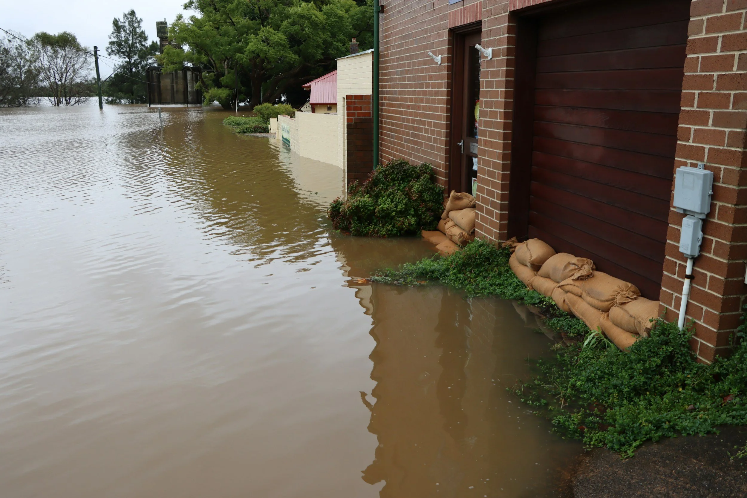 Floodwaters surround a brick building, with sandbags placed at the base of the wall to prevent further water entry. The water level is high, reaching the door and partially submerging the sidewalk.
