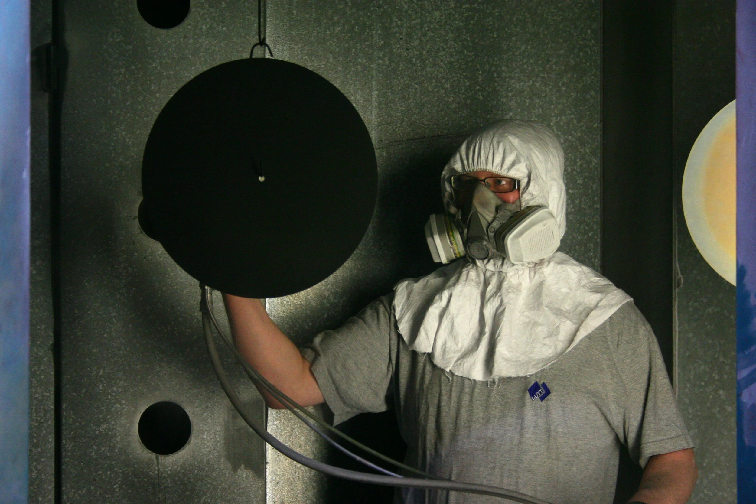 Person wearing a protective suit, mask, and glasses holding a black circular object in an industrial or laboratory setting.