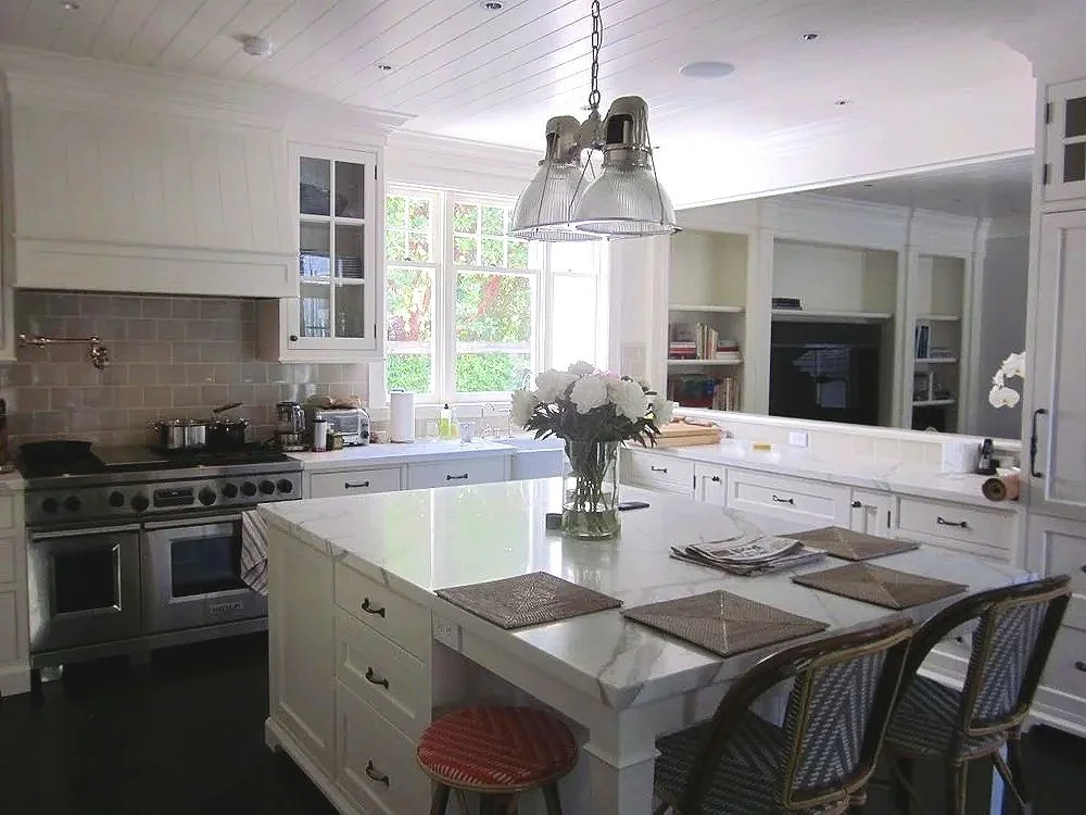 A bright, modern kitchen with white cabinets and a large island. The kitchen includes stainless steel appliances, a window with sunlight, a bouquet of white flowers on the island, and four upholstered chairs at the island. There are shelves with book